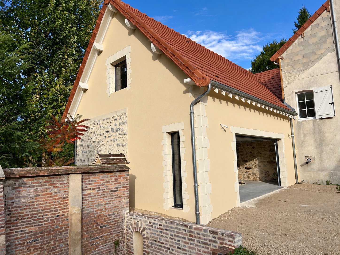 Bâtiment rénové à façade crépie beige, encadrements en pierre claire, toit en tuiles rouges et grande porte de garage, devant un muret de briques.