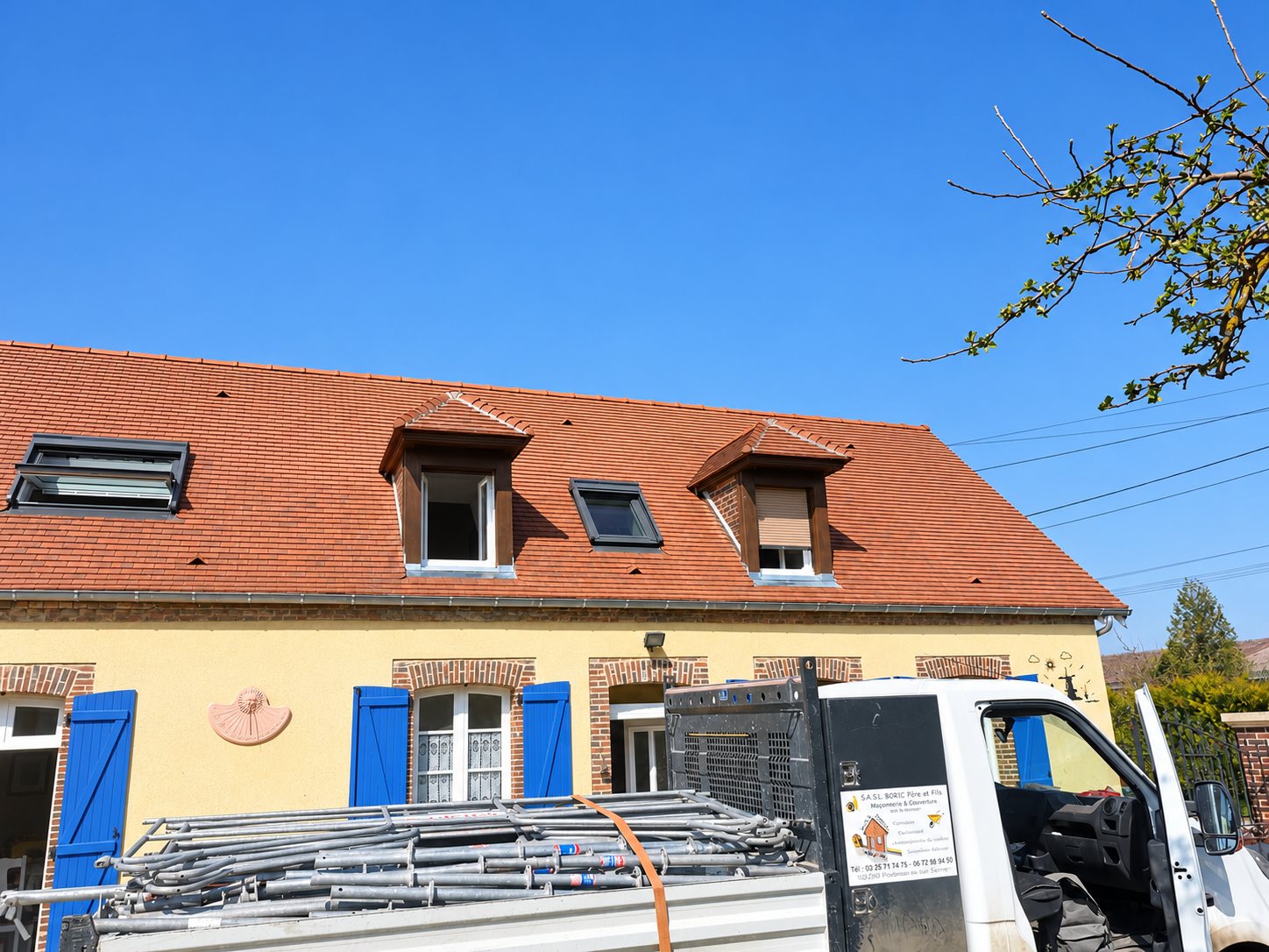 Un camion blanc chargé d'échafaudages est garé devant une maison jaune aux volets bleus, dont le toit de tuiles est équipé de lucarnes et de fenêtres de toit.