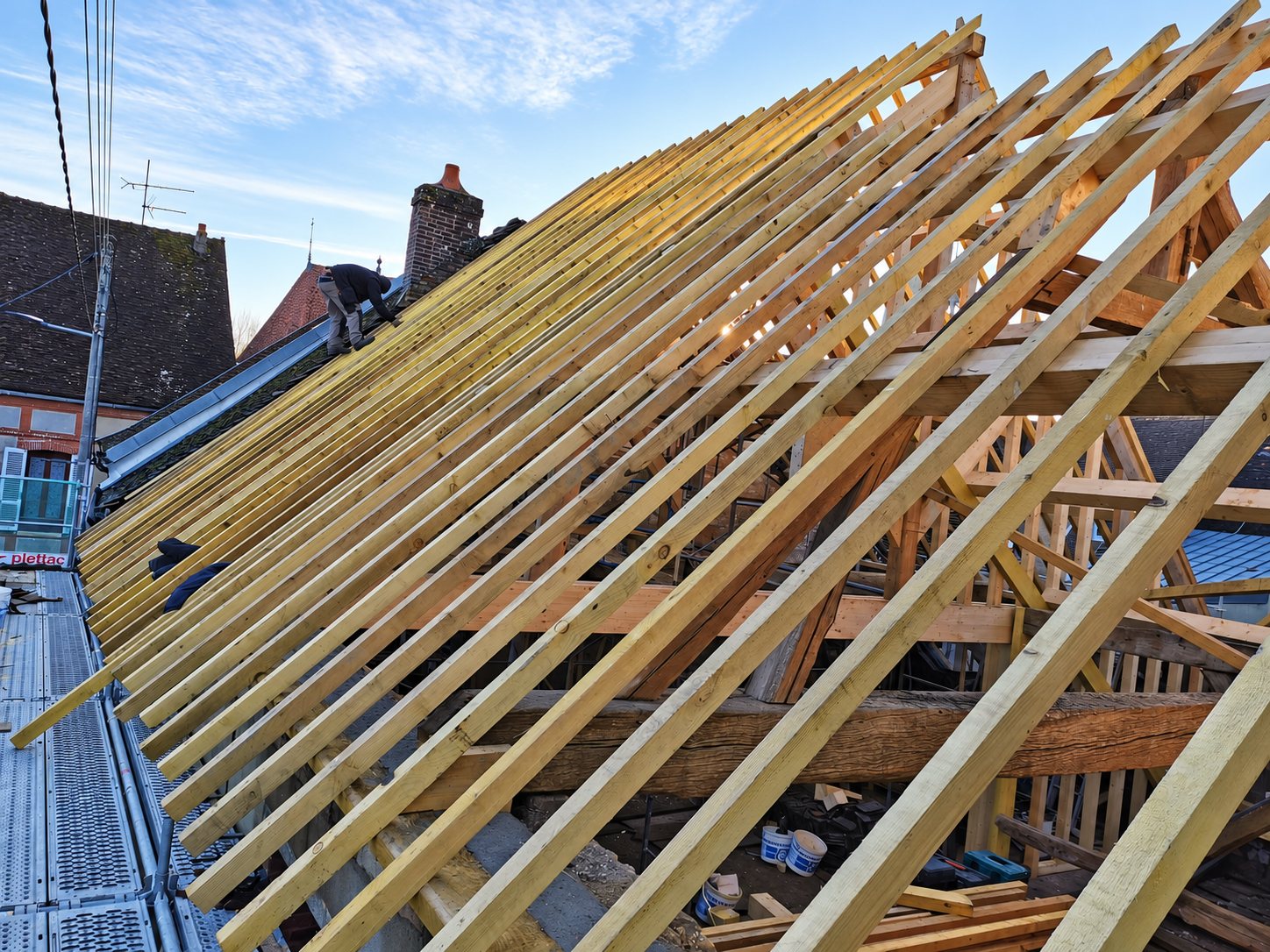 Une charpente ancienne en cours de rénovation, couverte de chevrons en bois neufs, avec un ouvrier travaillant sur le toit au coucher du soleil.