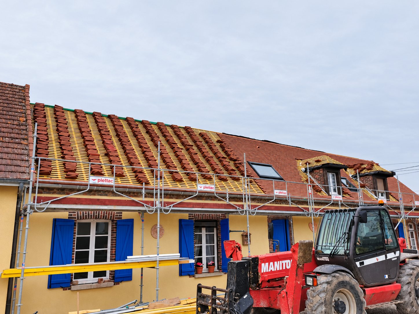 Une toiture en cours de rénovation, avec des tuiles posées sur les lattes, un échafaudage devant la façade jaune aux volets bleus et un chariot télescopique Manitou rouge au premier plan.