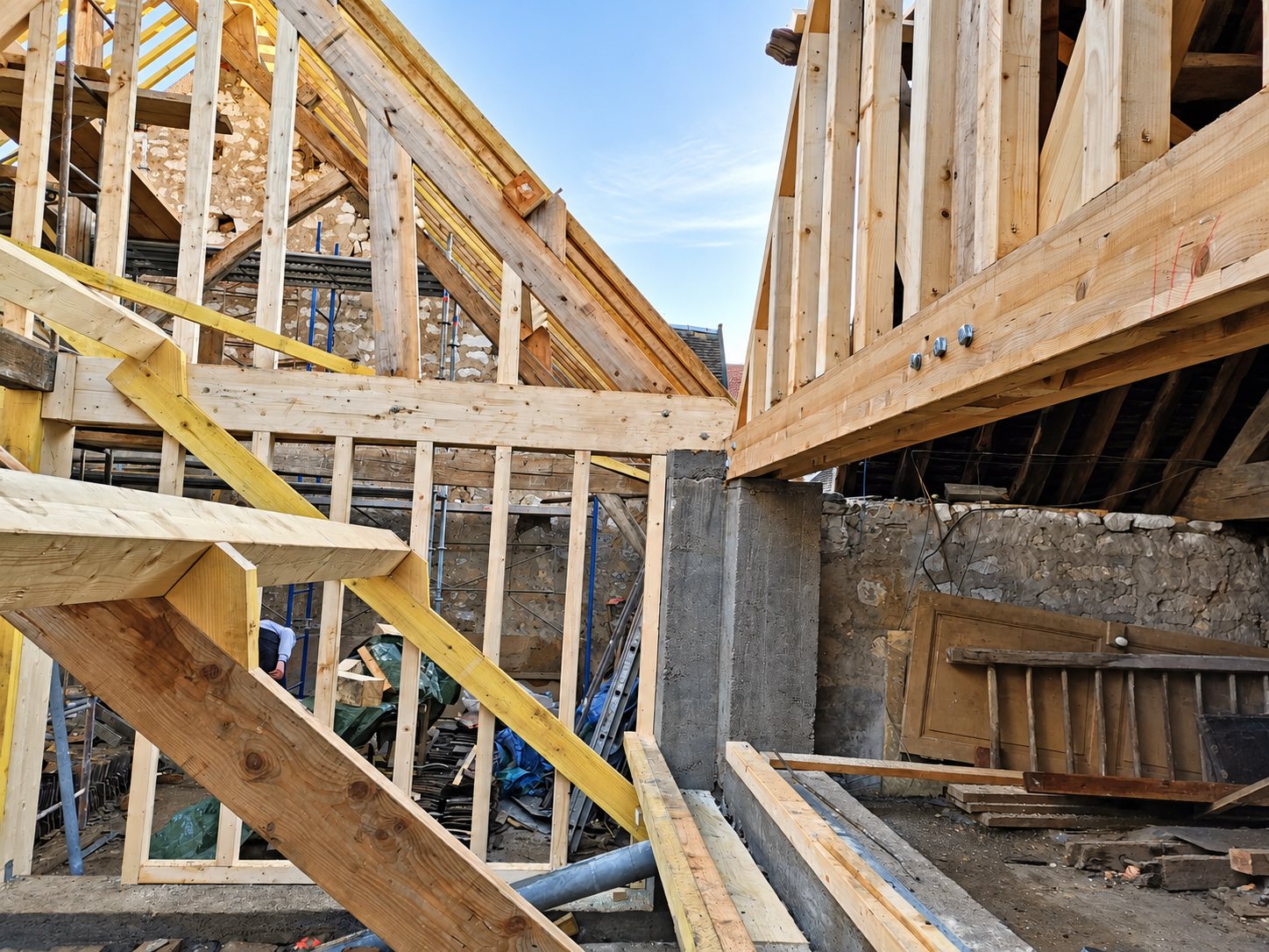 Chantier de rénovation montrant une charpente en bois neuve, des poutres assemblées par boulons et des murs en pierre anciens sous un ciel dégagé.