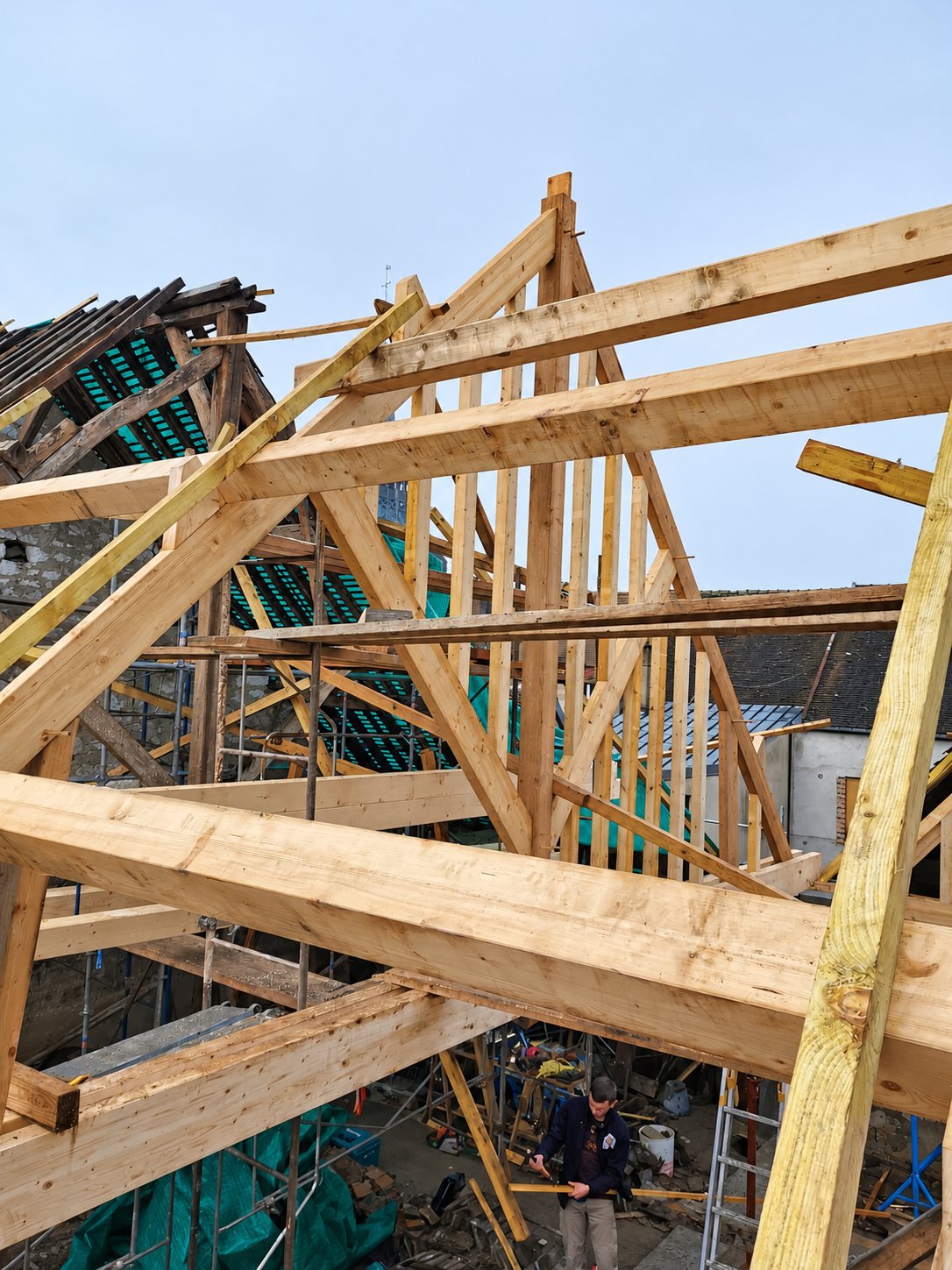 Charpente en bois neuve en cours de montage sur un bâtiment en pierre, avec échafaudages et un ouvrier au sol.