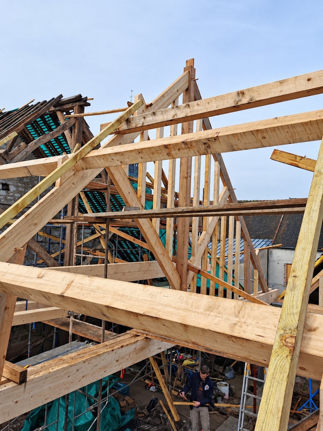 Structure de charpente en bois neuve montée sur un bâtiment en rénovation, avec échafaudages et un ouvrier au sol.