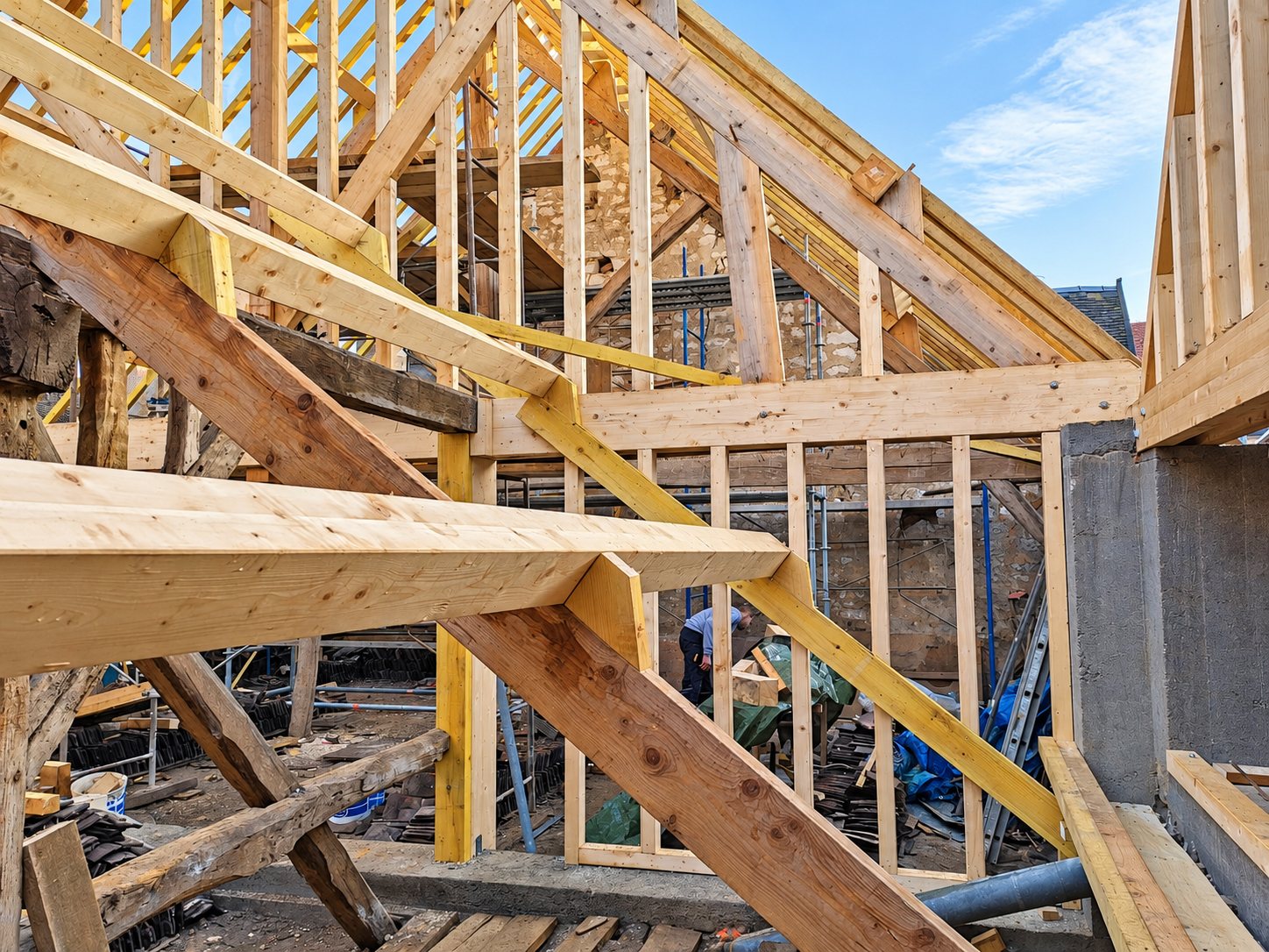 Structure de charpente en bois en cours de montage sur un chantier, avec des murs en pierre et un ouvrier au loin.