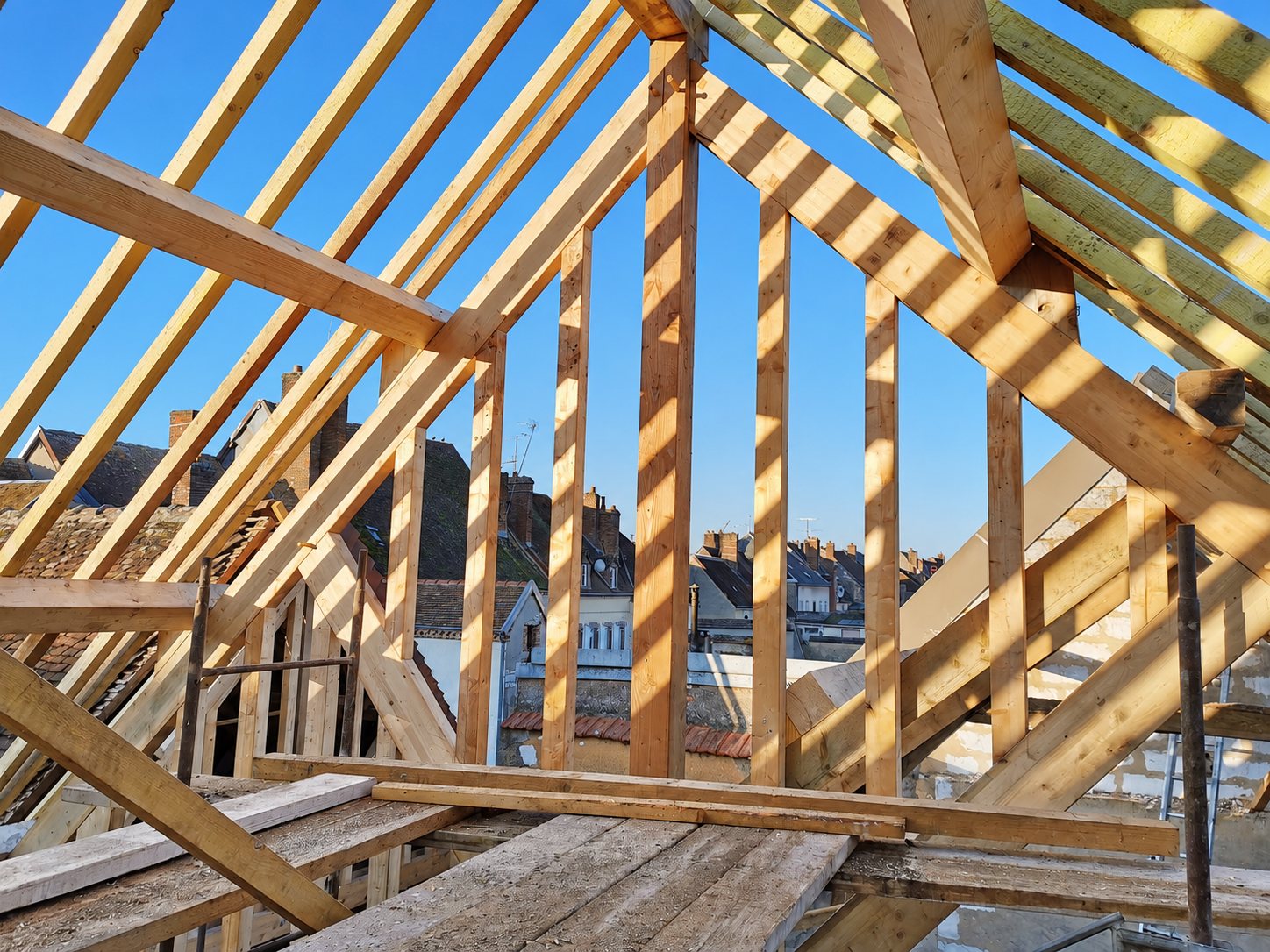 Une charpente traditionnelle en bois en cours de montage, éclairée par le soleil, avec des toits de maisons et un ciel bleu en arrière-plan.