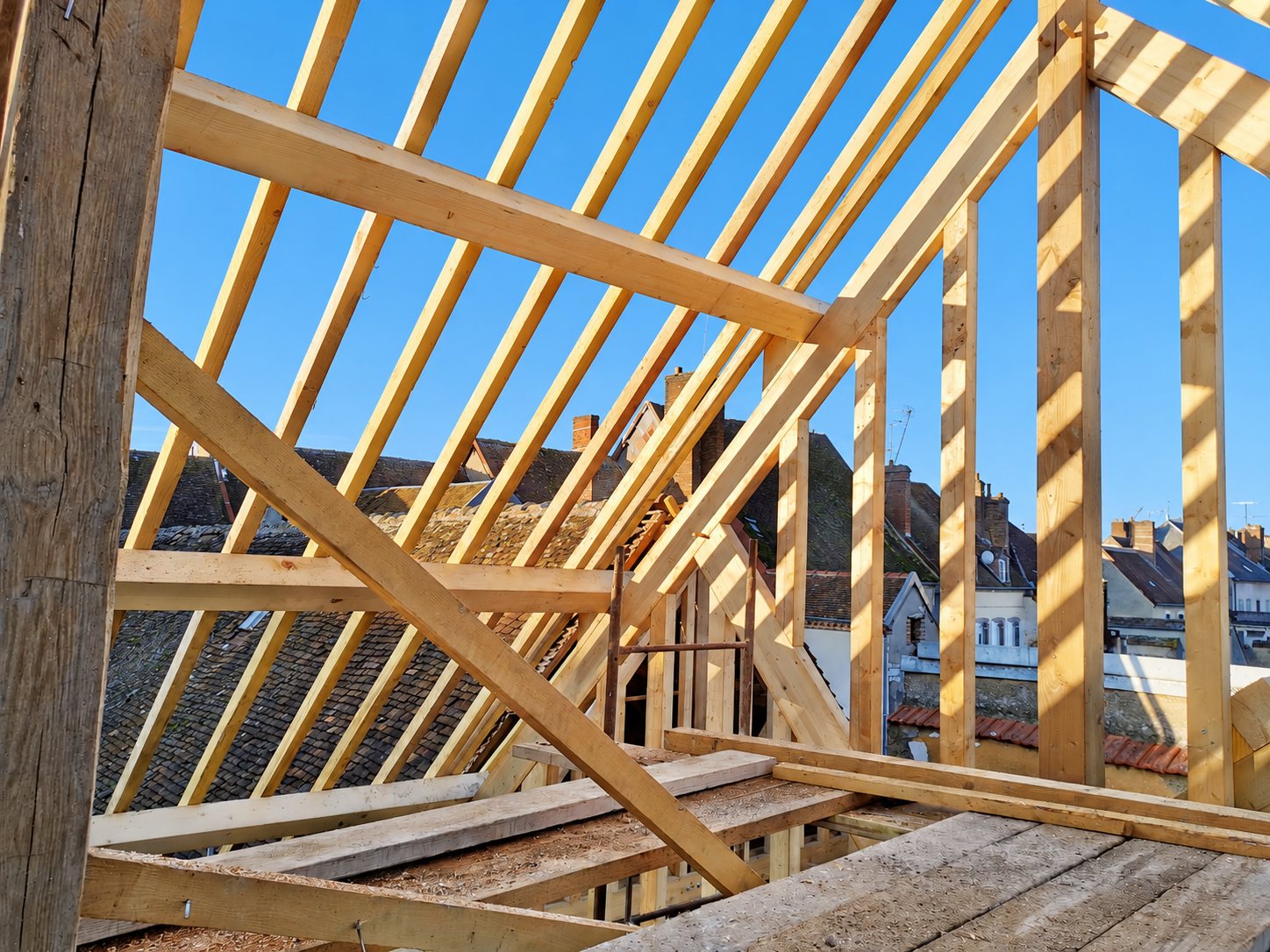 Une charpente en bois neuve en cours de montage, avec chevrons et poutres éclairés par le soleil, au-dessus des toits d'une ville sous un ciel bleu.