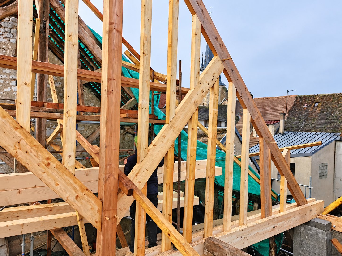 Une charpente en bois neuve en cours de montage sur un bâtiment en pierre, avec un ouvrier et des toits de village en arrière-plan.