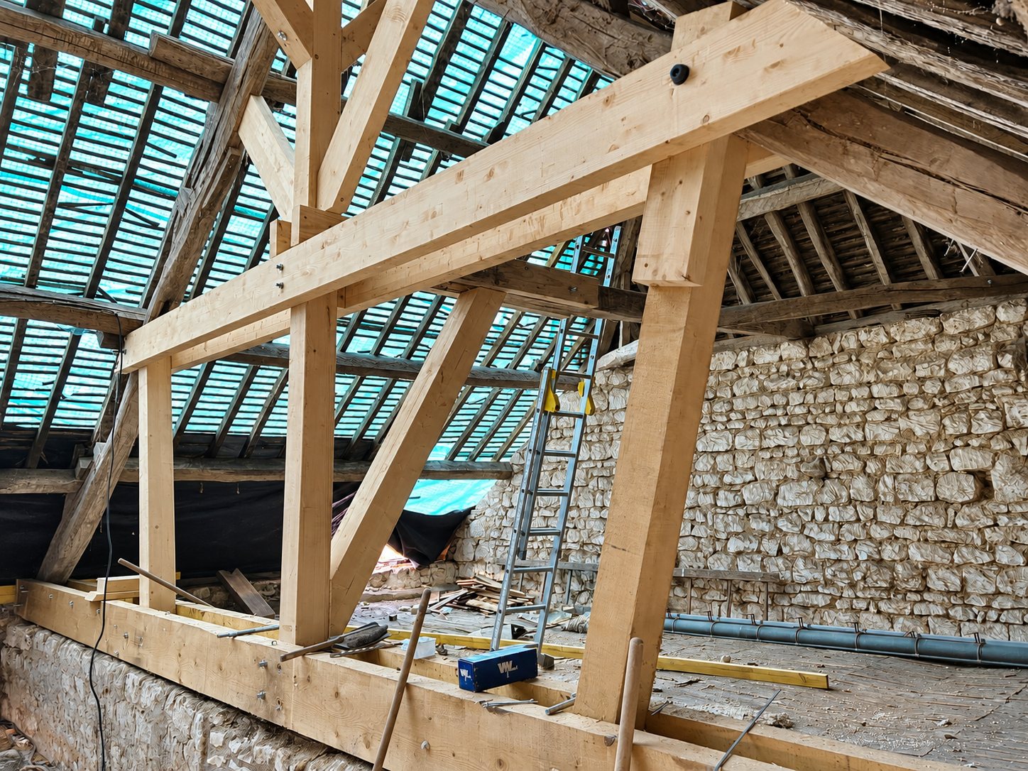 Charpente en bois neuve installée sous une toiture de tuiles anciennes, dans une grange aux murs de pierre en cours de rénovation.