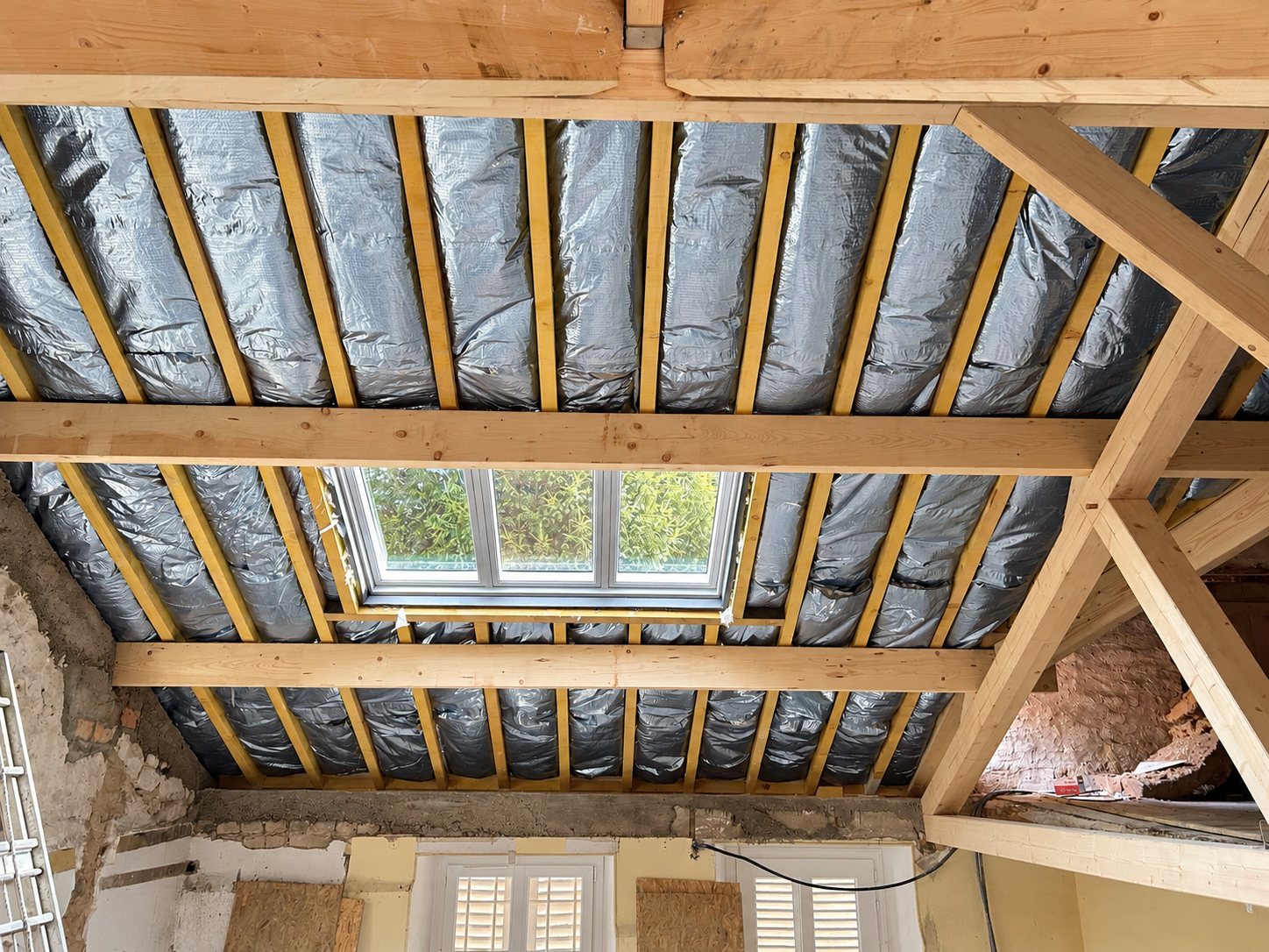 Vue de dessous d'une charpente en bois dont les chevrons sont garnis d'isolant réflecteur argenté, avec une fenêtre de toit au centre, dans un chantier de rénovation.