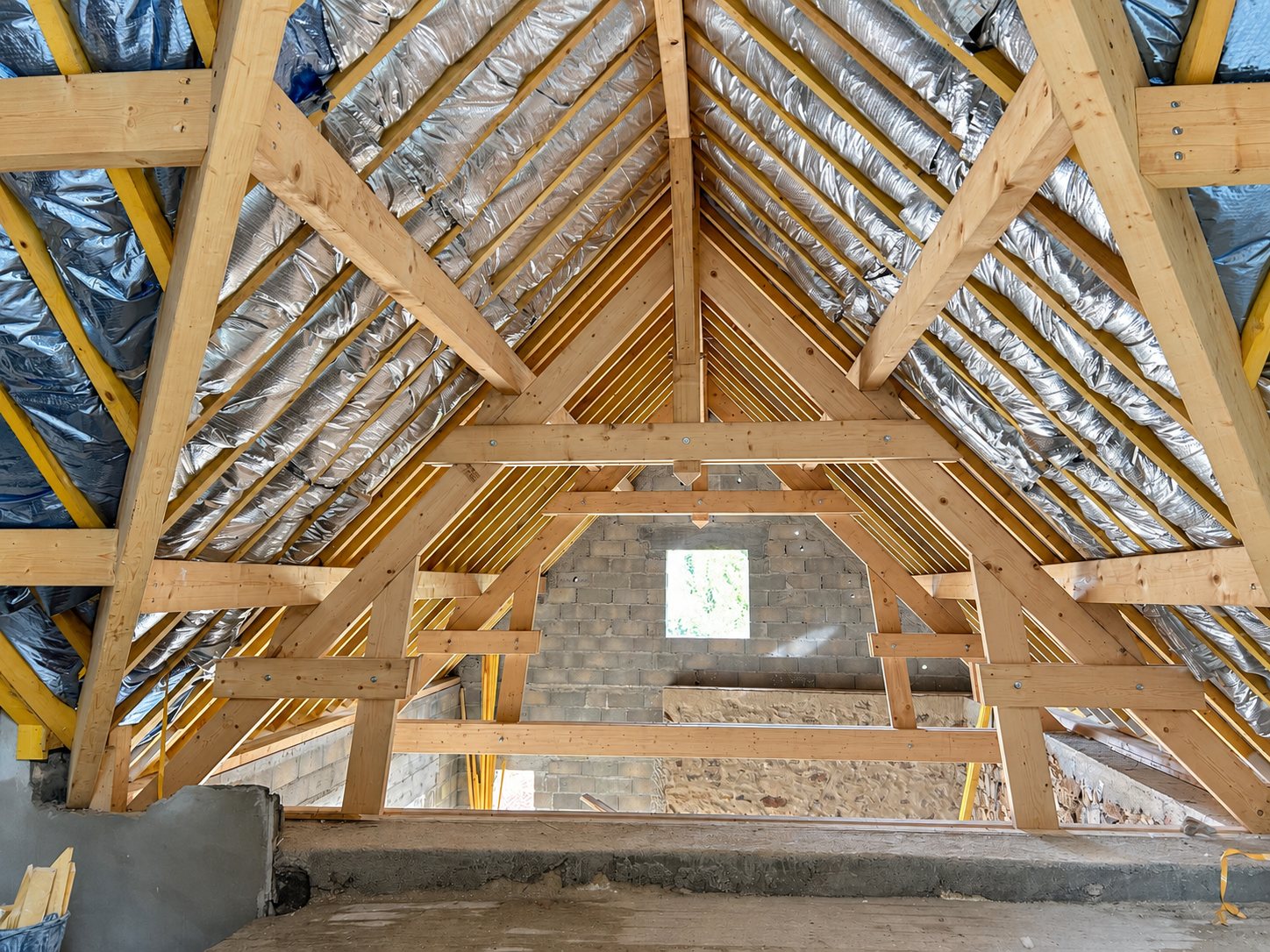 Vue intérieure d'une charpente traditionnelle en bois dont les chevrons sont recouverts d'un isolant réflecteur argenté, devant un mur en parpaings.