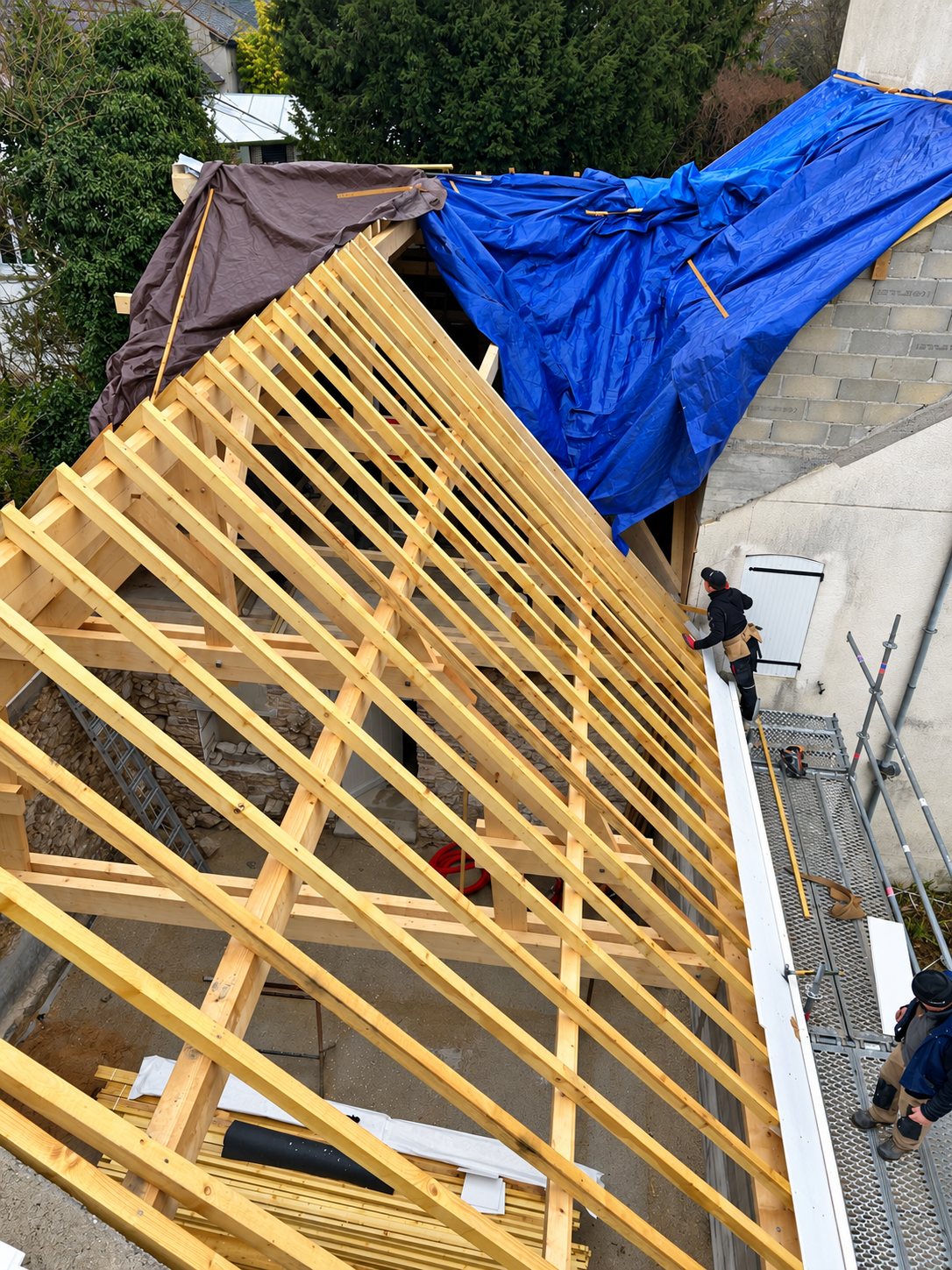 Vue en hauteur d'une charpente en bois en cours de pose, avec des bâches bleue et marron et des ouvriers sur un échafaudage à côté d'un mur en pierre.