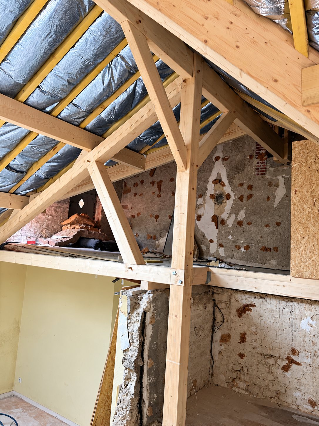 Combles en cours de rénovation avec une charpente en bois clair, un isolant réfléchissant argenté sous la toiture et des murs anciens en pierre et en torchis.
