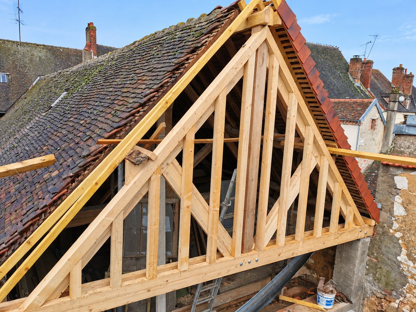 Une charpente en bois clair en cours de construction sur un pignon, à côté d'un ancien toit de tuiles plates moussues et de maisons de village.
