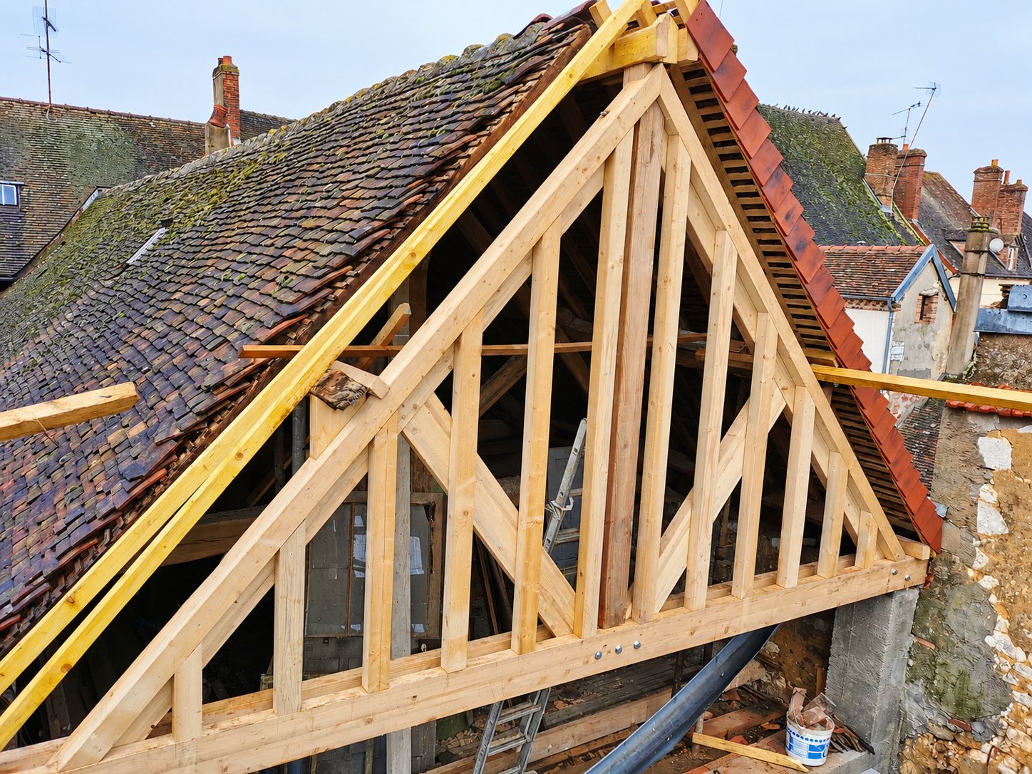 Une charpente traditionnelle en bois clair en cours de construction sur le pignon d'une vieille maison au toit de petites tuiles brunes.