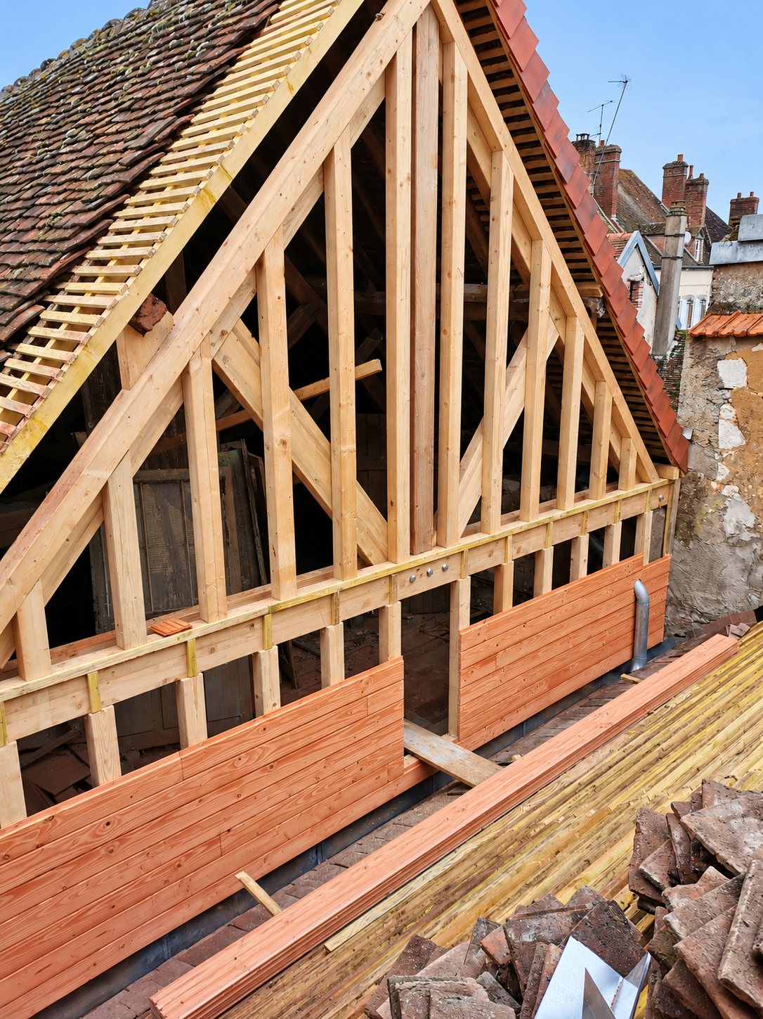 Ossature en bois neuve d'un pignon de toit, avec bardage en cours de pose et tuiles anciennes empilées sur un chantier de rénovation.