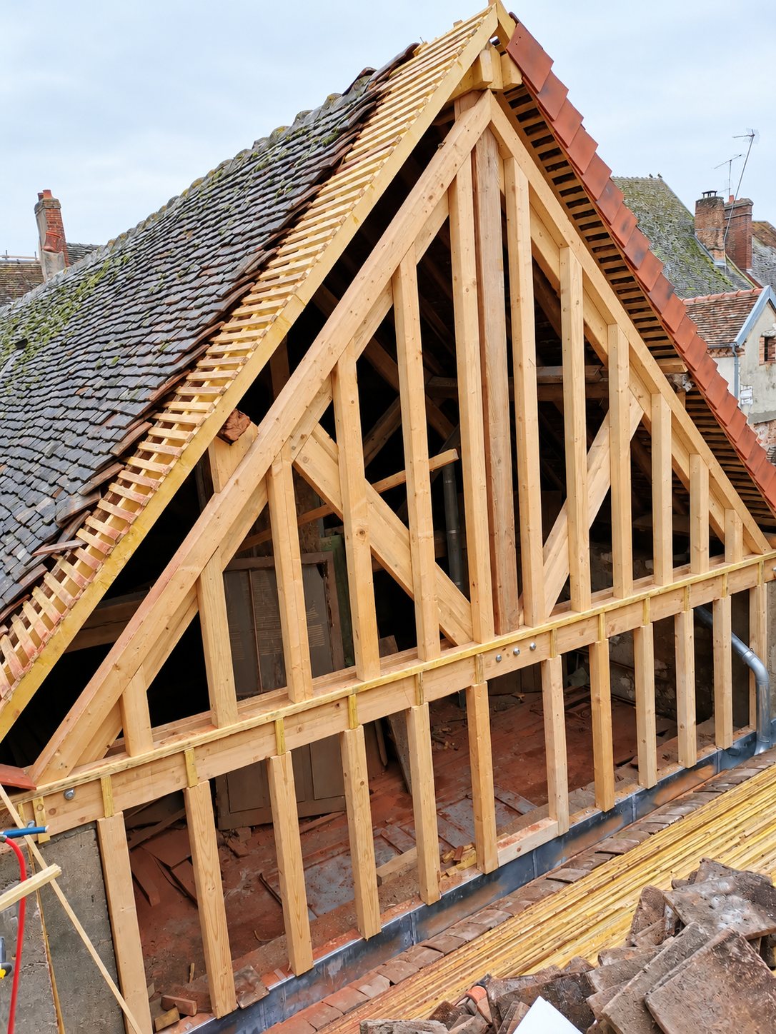 Une charpente en bois clair formant un pignon triangulaire, avec des tuiles anciennes empilées au premier plan, sur un chantier de rénovation de toiture.