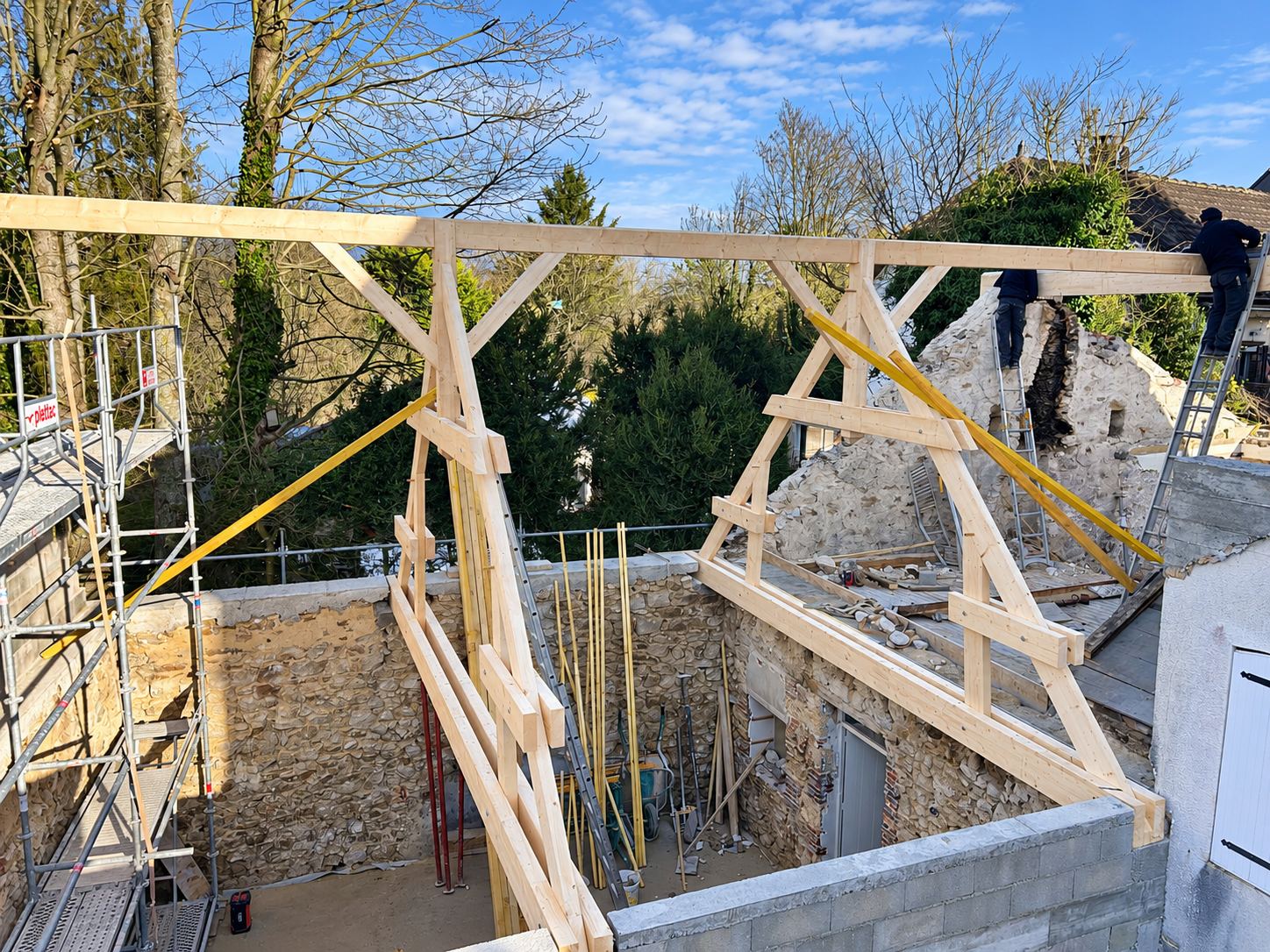 Des charpentiers installent une ossature de charpente en bois clair sur les murs en pierre d'un bâtiment en rénovation, avec un échafaudage à gauche.