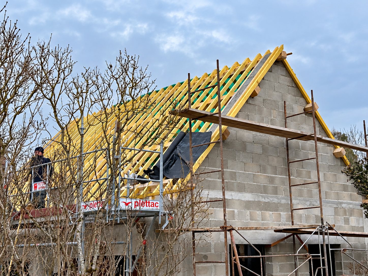 Une charpente en bois clair est en cours de montage sur une maison en parpaings entourée d'échafaudages, avec un ouvrier sur une plateforme à gauche.