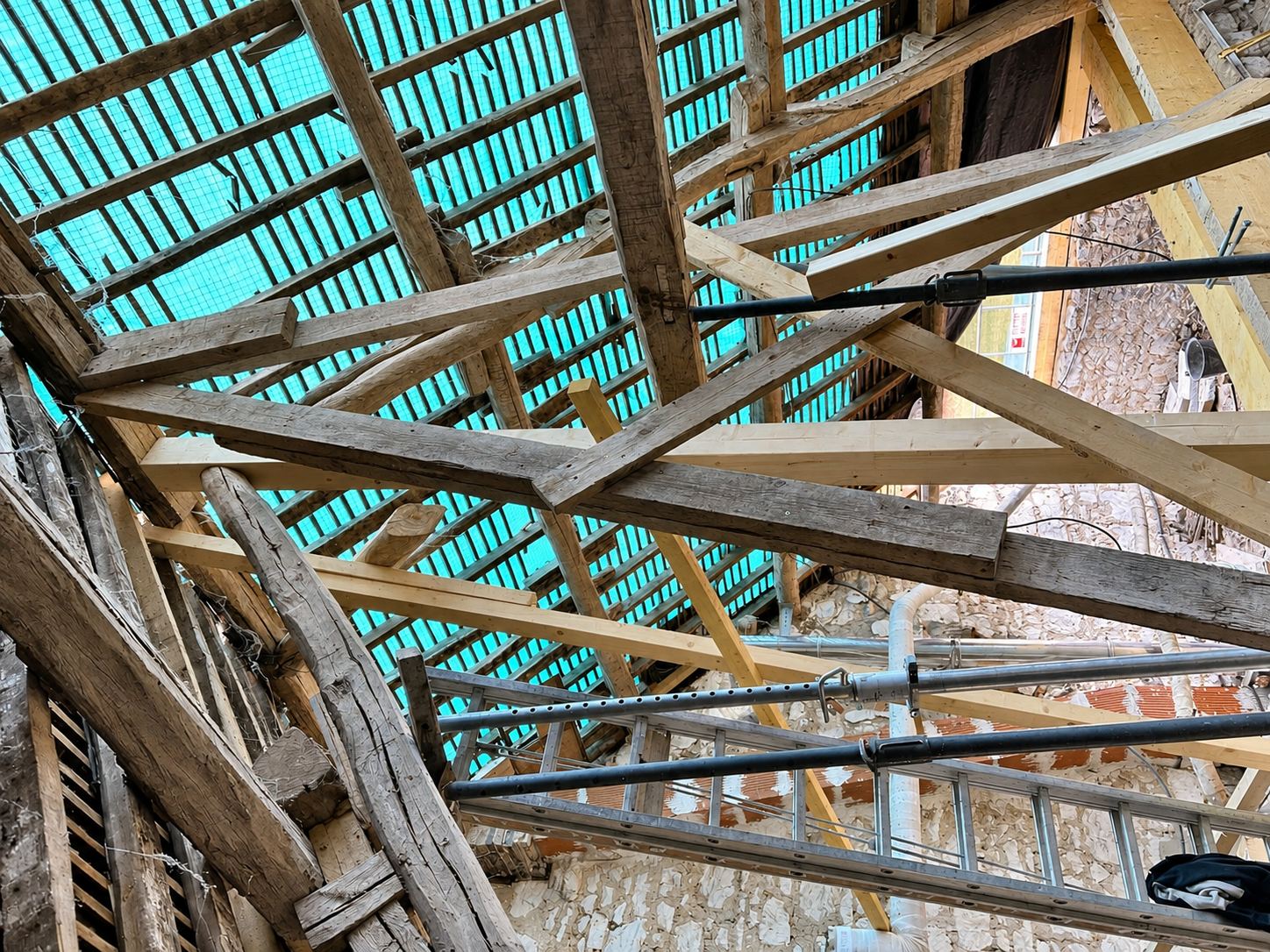 Vue depuis l'intérieur d'une vieille charpente en bois en cours de rénovation, recouverte d'une bâche turquoise et soutenue par des étais métalliques devant un mur en pierre.