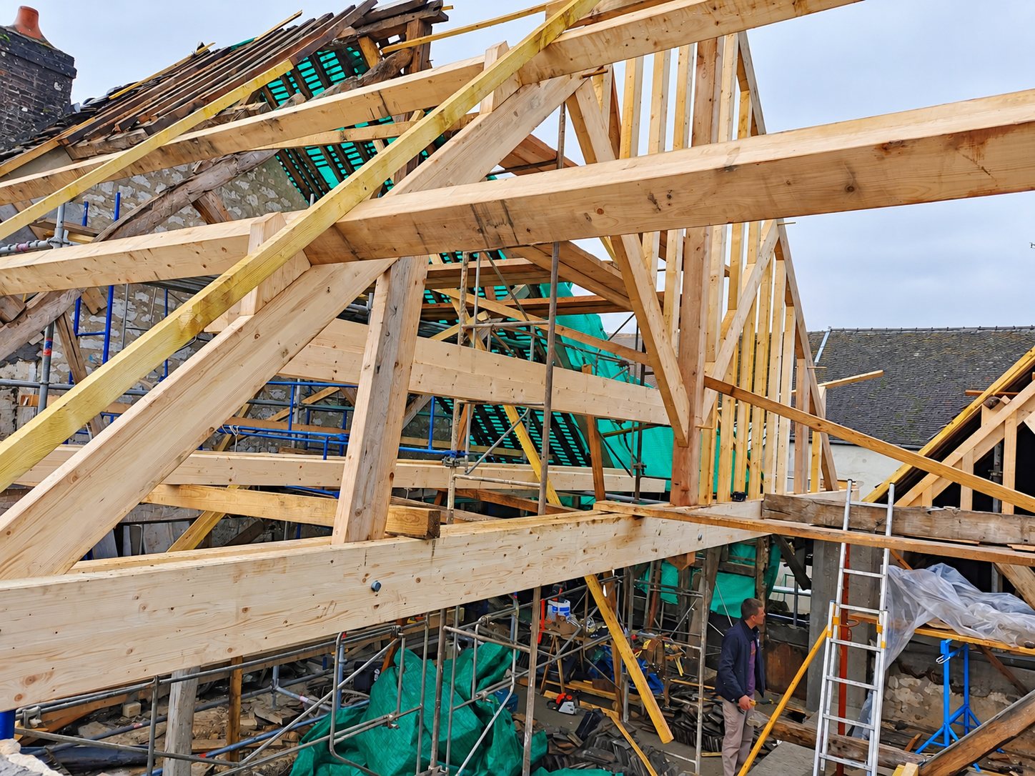 Une charpente en bois neuve en cours de montage sur un bâtiment en pierre, entourée d'échafaudages et de bâches vertes, avec un ouvrier au fond.