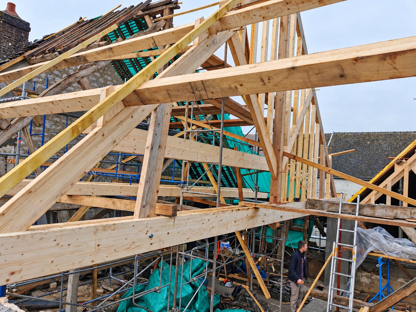 Une charpente en bois neuve en cours de montage sur un bâtiment en pierre, avec échafaudages, bâches vertes et un ouvrier au sol.