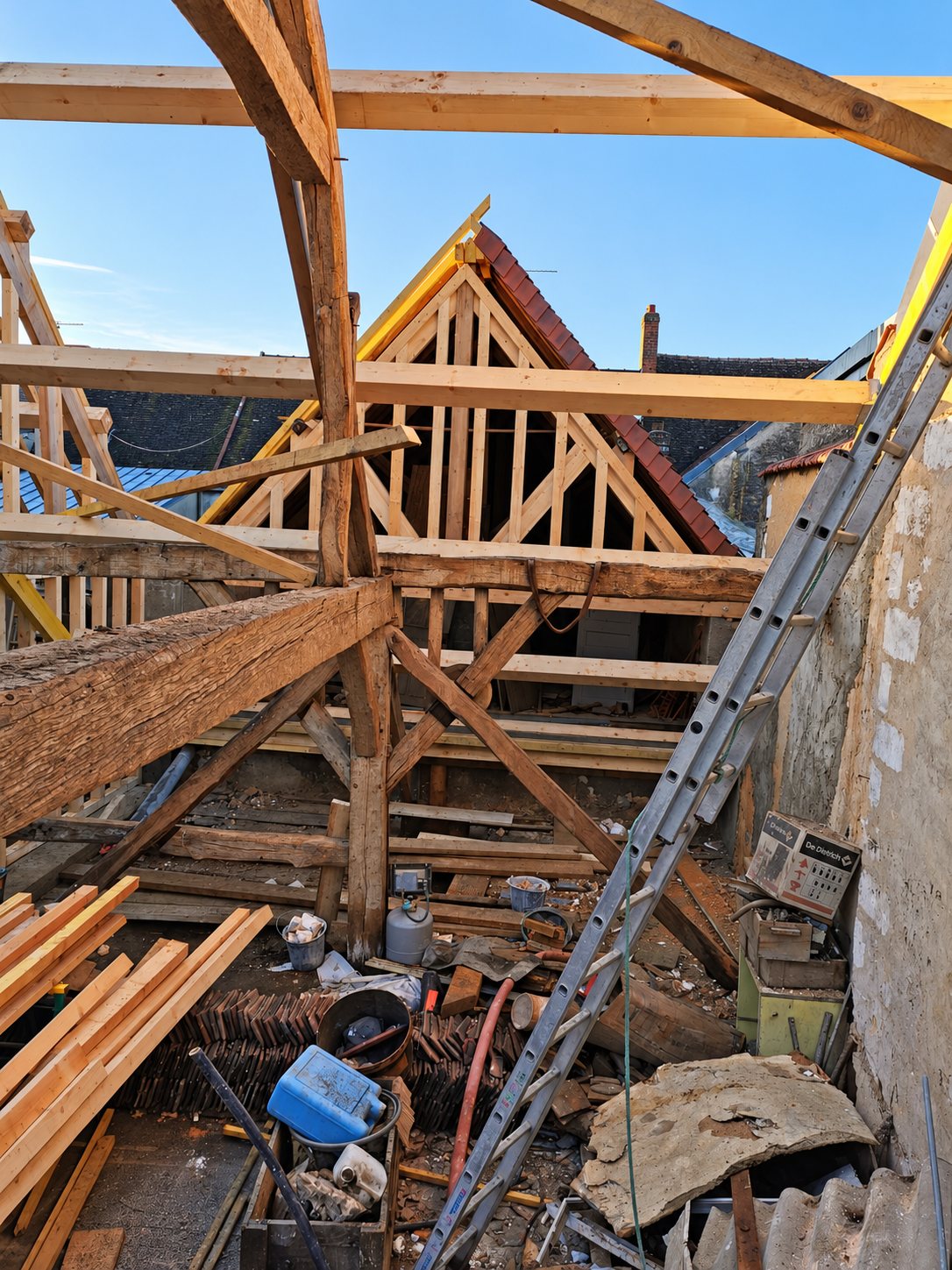 Chantier de rénovation d'une charpente traditionnelle en bois avec poutres anciennes, planches neuves, échelle et gravats au sol sous un ciel dégagé.