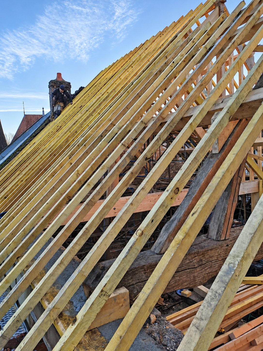 Des chevrons en bois clair recouvrent une charpente ancienne en chantier, tandis qu'un ouvrier travaille sur le toit voisin sous un ciel dégagé.