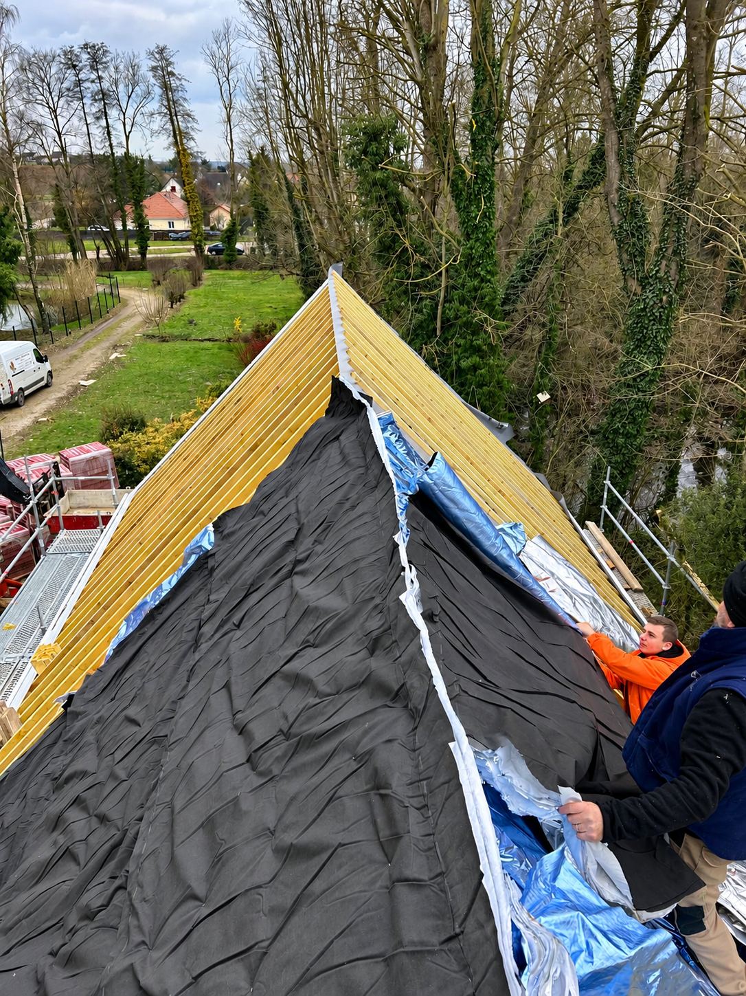 Deux couvreurs déroulent un écran de sous-toiture noir et isolant argenté sur une charpente en bois, avec des tuiles stockées sur un échafaudage et des arbres autour.