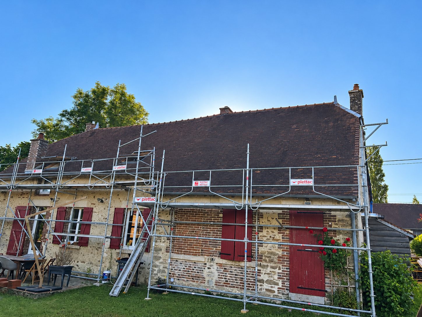 Une maison ancienne en pierre et brique aux volets rouges est entourée d'un échafaudage métallique plettac, sous un ciel bleu dégagé.