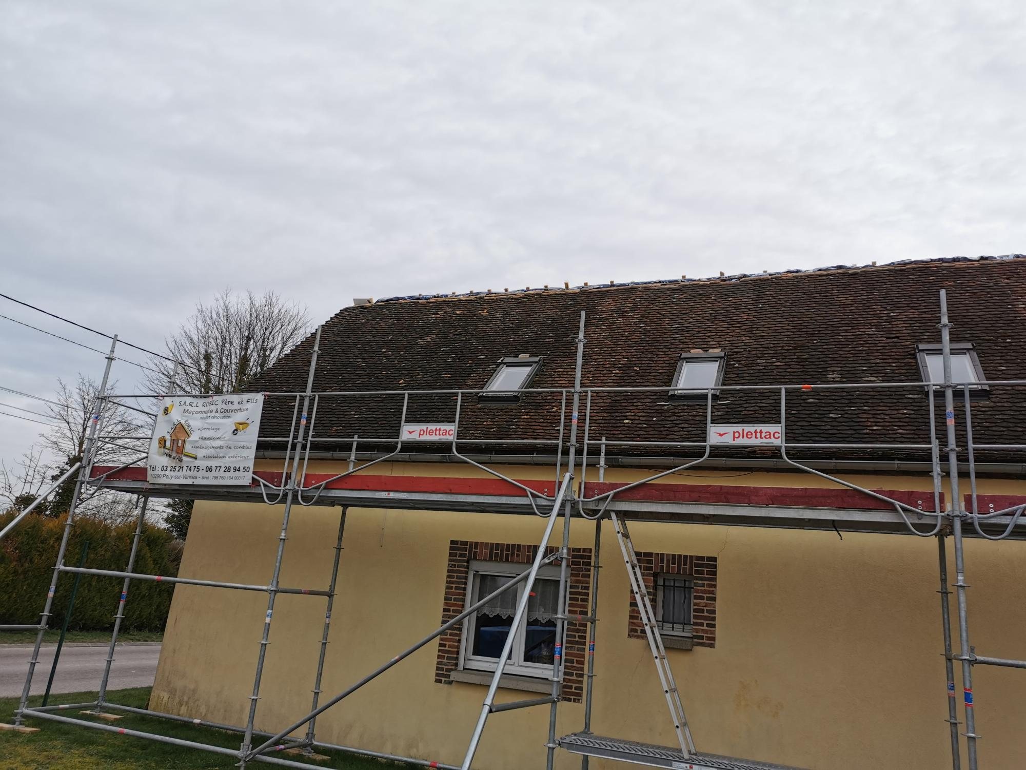 Une maison jaune à toit de tuiles brunes entourée d'un échafaudage plettac avec une bâche publicitaire d'entreprise de maçonnerie et couverture.