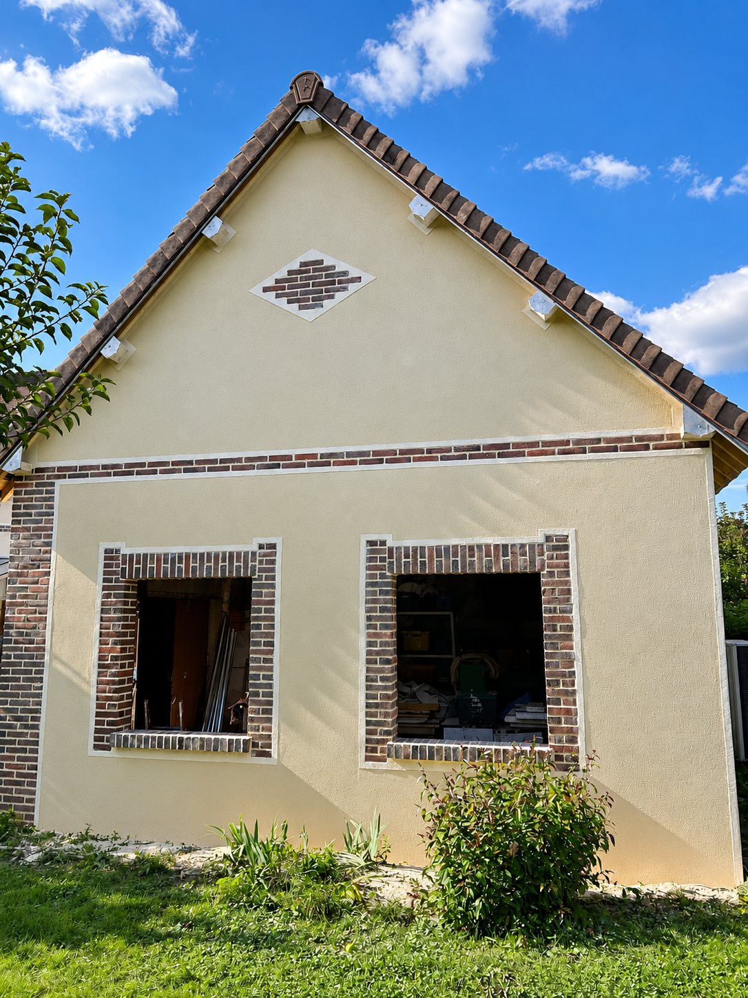 Pignon d'une maison fraîchement ravalé avec un enduit beige, des encadrements de fenêtres en briques rouges et un motif en losange sous le toit.
