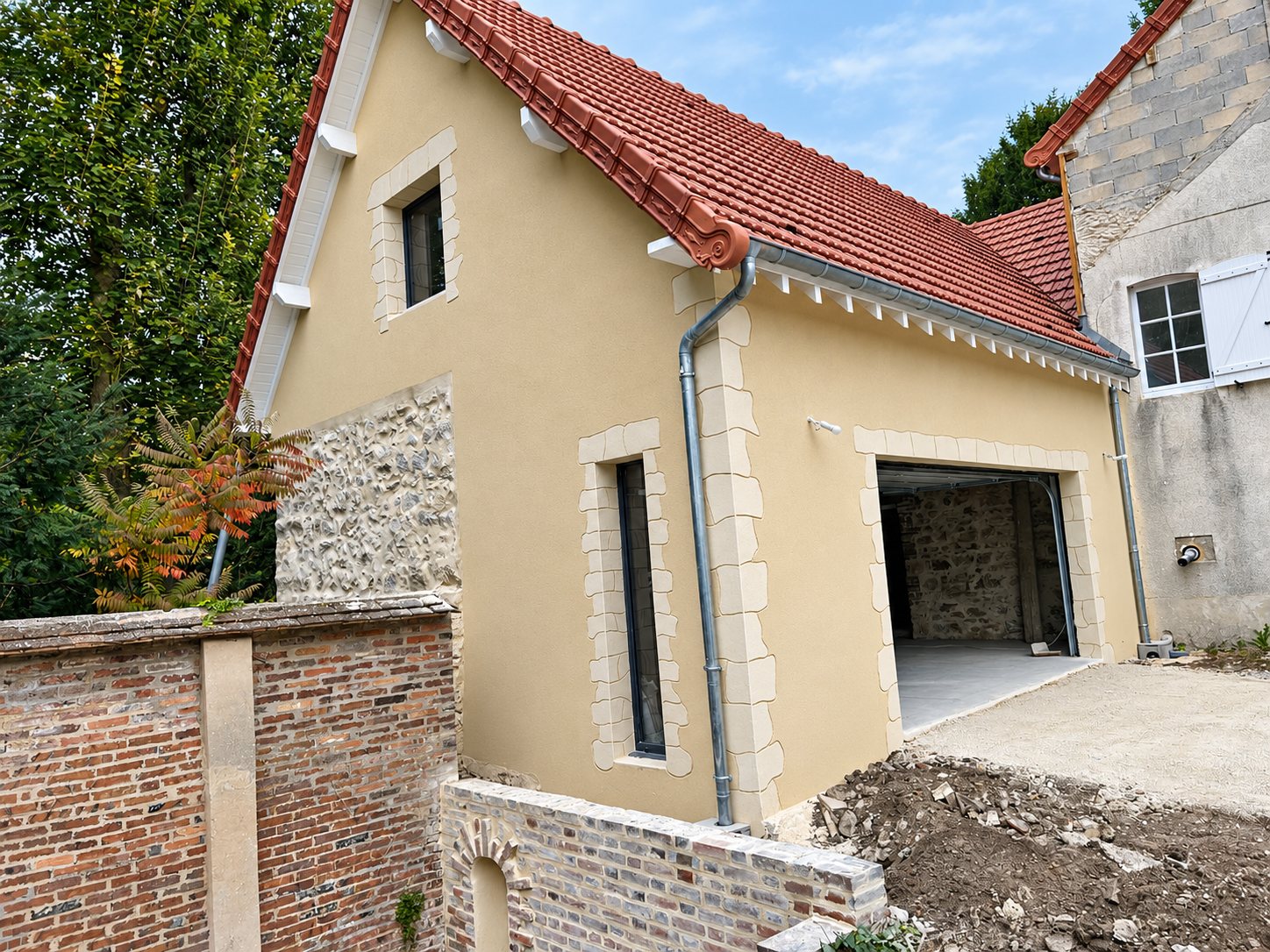 Façade d'une maison récemment rénovée avec enduit beige, encadrements en pierre, toit en tuiles rouges et garage ouvert, près d'un mur en briques.