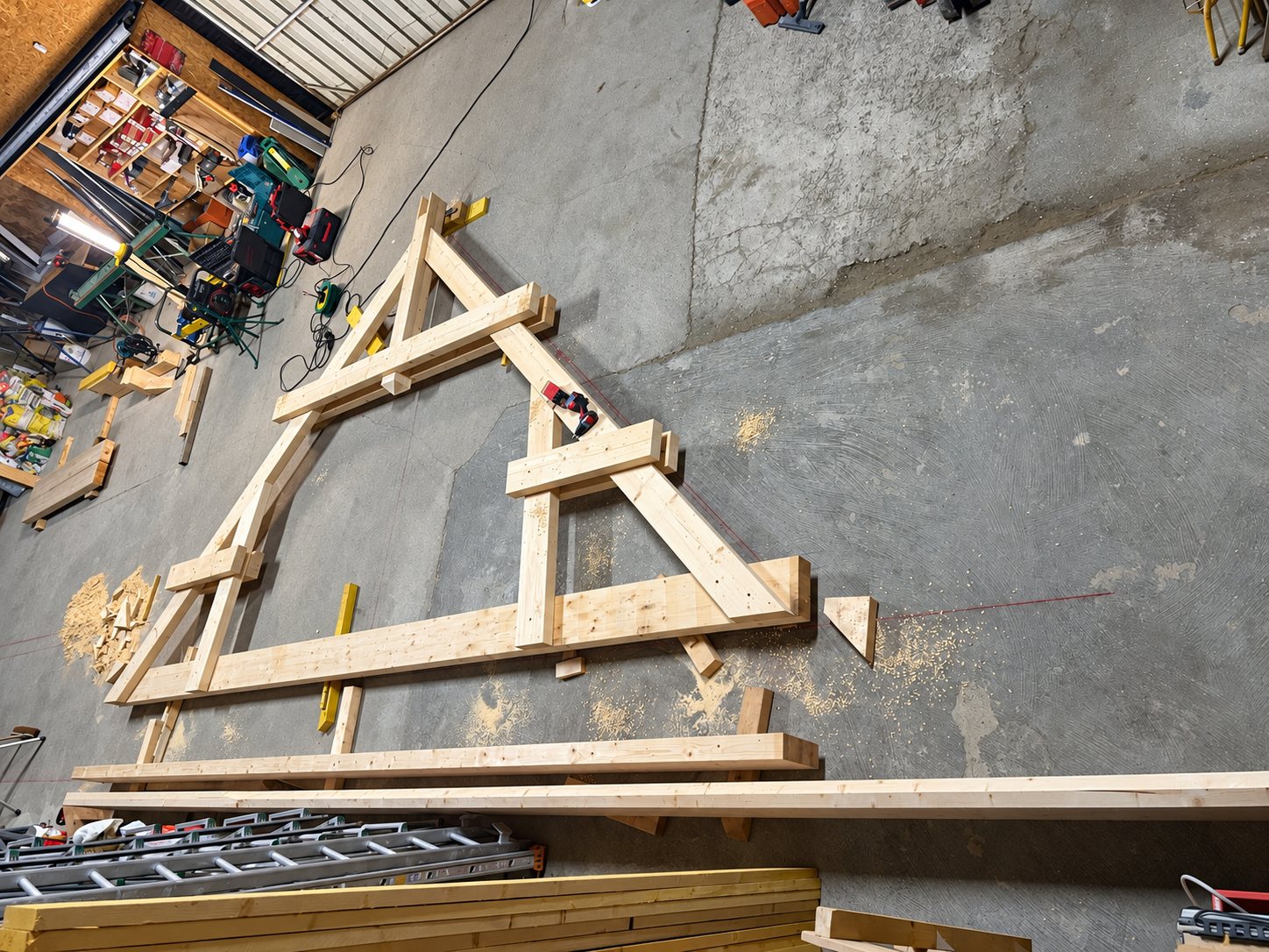 Une ferme de charpente en bois clair est assemblée à plat sur le sol en béton d'un atelier, entourée de sciure, d'outils et de planches.