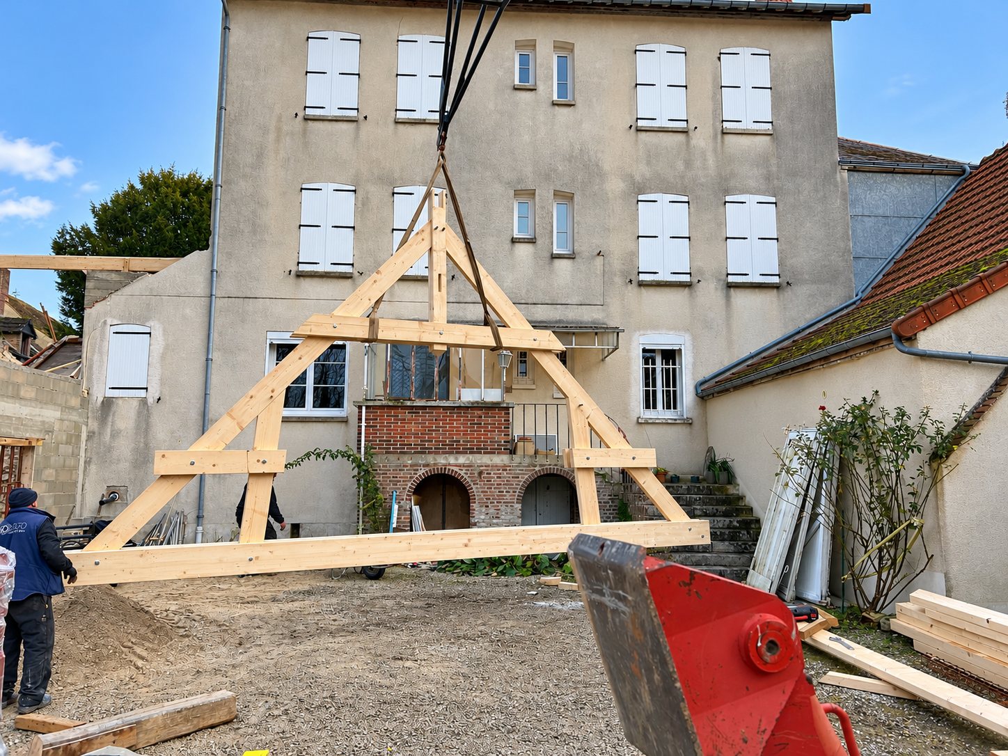 Une ferme de charpente en bois claire est suspendue à une grue dans une cour, devant une grande maison en pierre aux volets blancs.