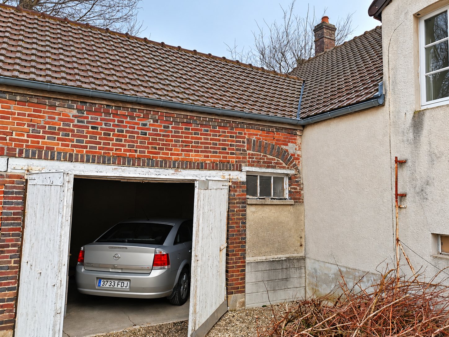 Une voiture grise est garée dans un garage en brique rouge aux portes blanches ouvertes, sous un toit de tuiles brunes, à côté d'une maison crépie.