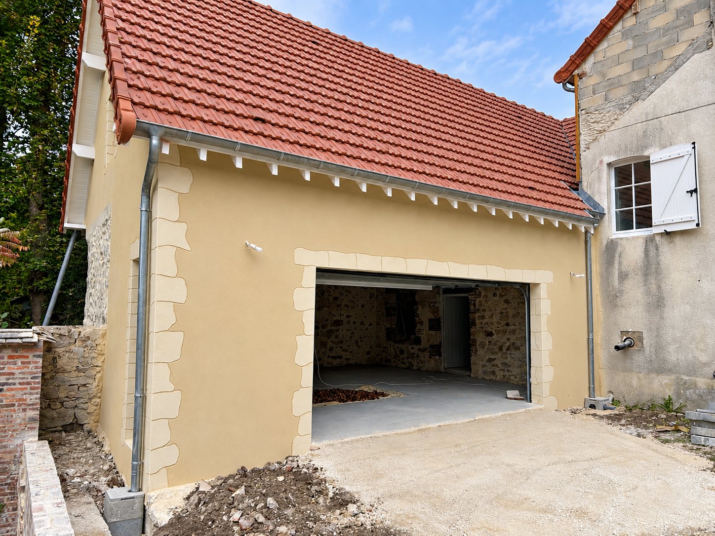 Façade d'un garage rénové avec enduit beige, encadrements en pierre et toit en tuiles rouges, devant une allée de gravier.