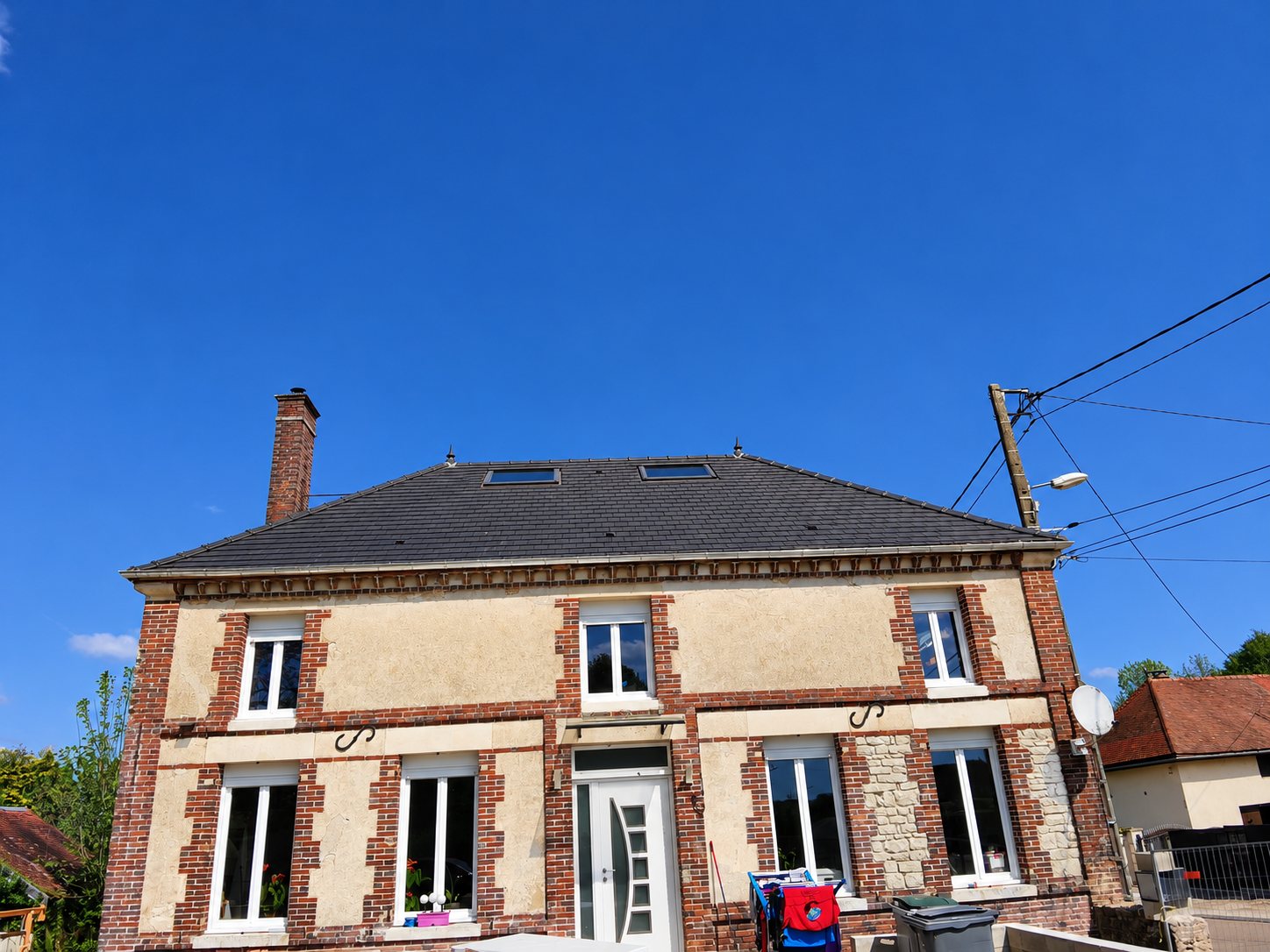 Façade d'une maison ancienne en briques rouges et pierre claire, dotée d'un toit sombre équipé de deux fenêtres de toit, sous un ciel bleu.