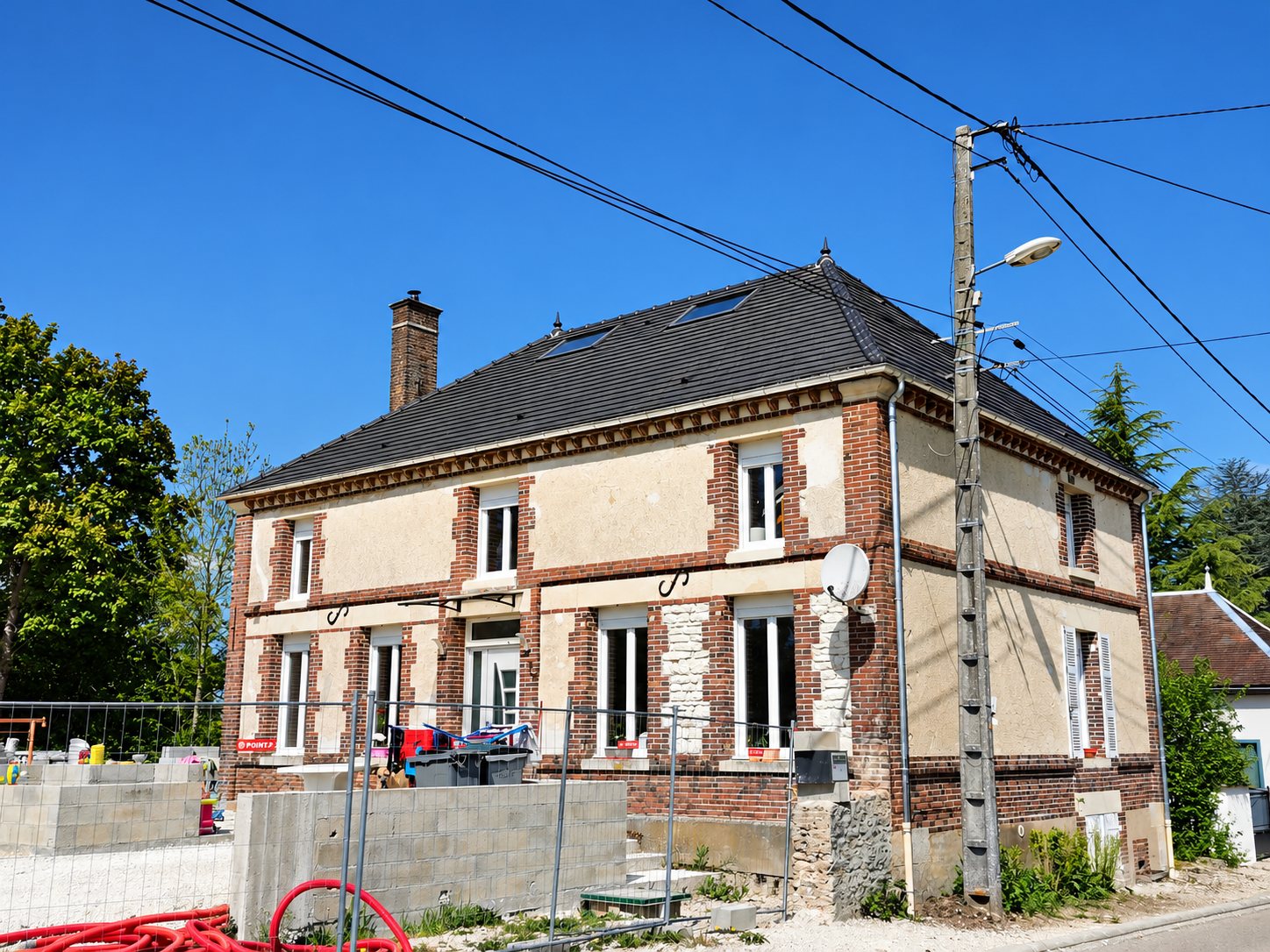 Maison ancienne en brique et enduit clair dotée d'une toiture en ardoise neuve avec fenêtres de toit, devant un chantier clôturé avec parpaings et gaine rouge.