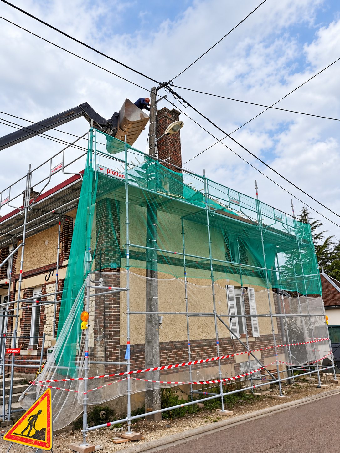 Une maison en briques recouverte d'échafaudages et de filets verts, avec un ouvrier près de la toiture et un panneau de travaux au premier plan.