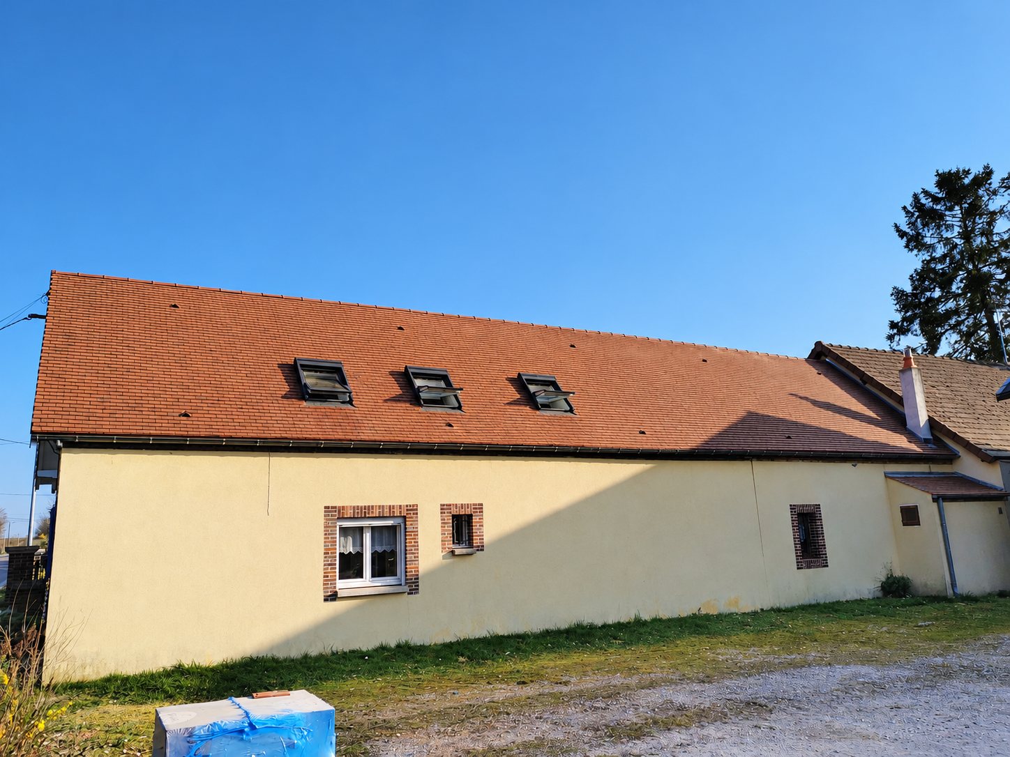 Façade d'une maison aux murs jaunes surmontée d'un toit en tuiles rouges équipé de trois fenêtres de toit, sous un ciel bleu.