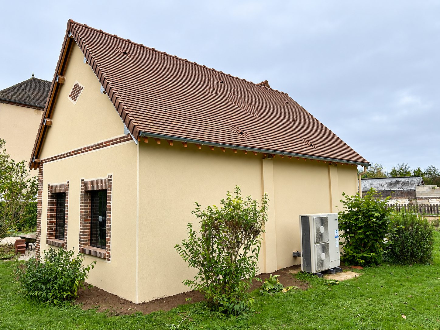 Petite maison à la façade enduite beige et au toit de tuiles brunes, avec une pompe à chaleur installée contre le mur et entourée d'un jardin.