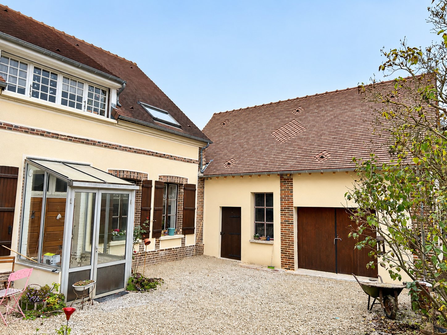 Cour gravillonnée entourée d'une maison aux façades crème et briques avec véranda, et d'une grange à la toiture en tuiles neuves et aux portes de bois brun.