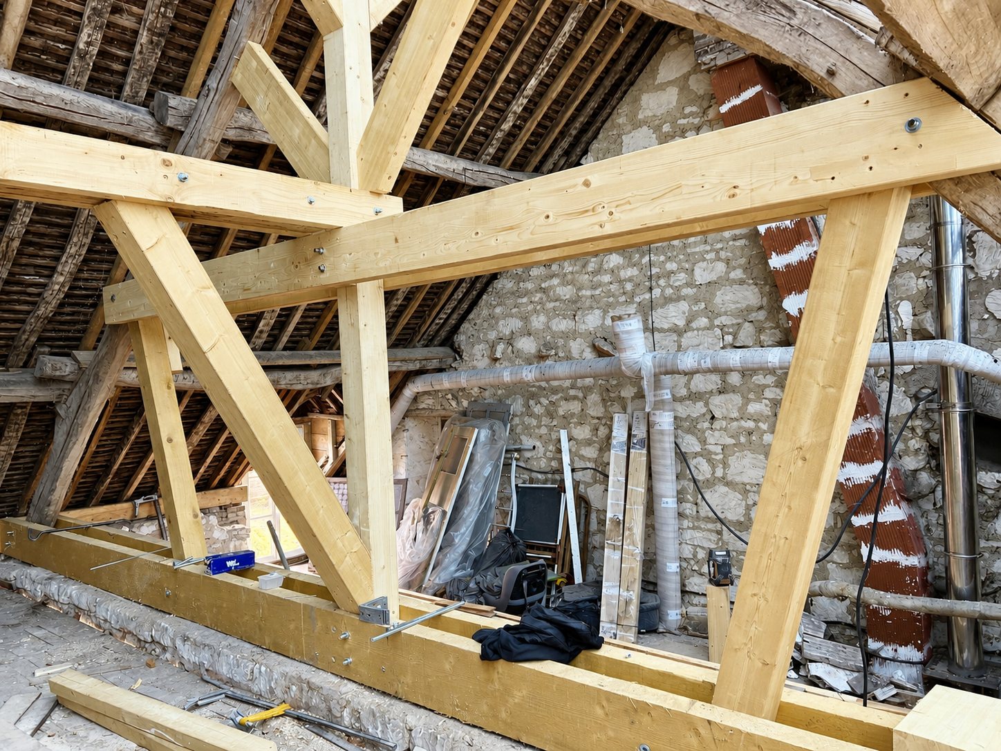 Charpente neuve en bois clair installée sous d'anciennes tuiles dans un grenier aux murs de pierre en cours de rénovation.