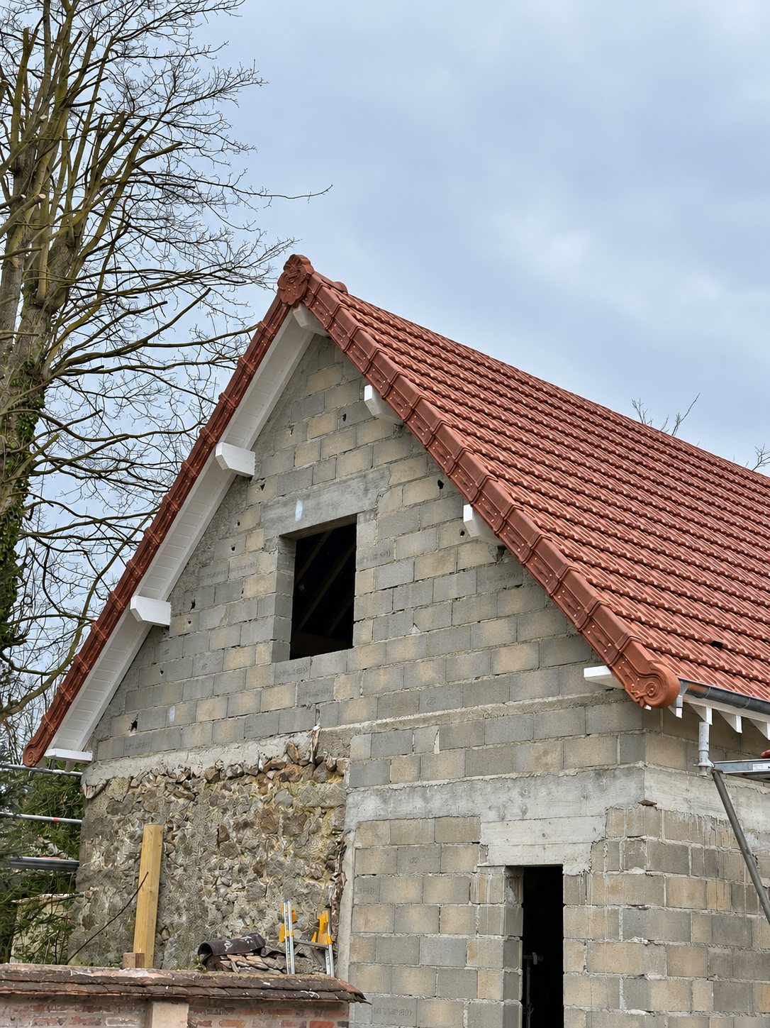 Pignon d'un bâtiment en parpaings et pierres surmonté d'une toiture neuve en tuiles rouges avec rives décoratives et chevrons blancs.