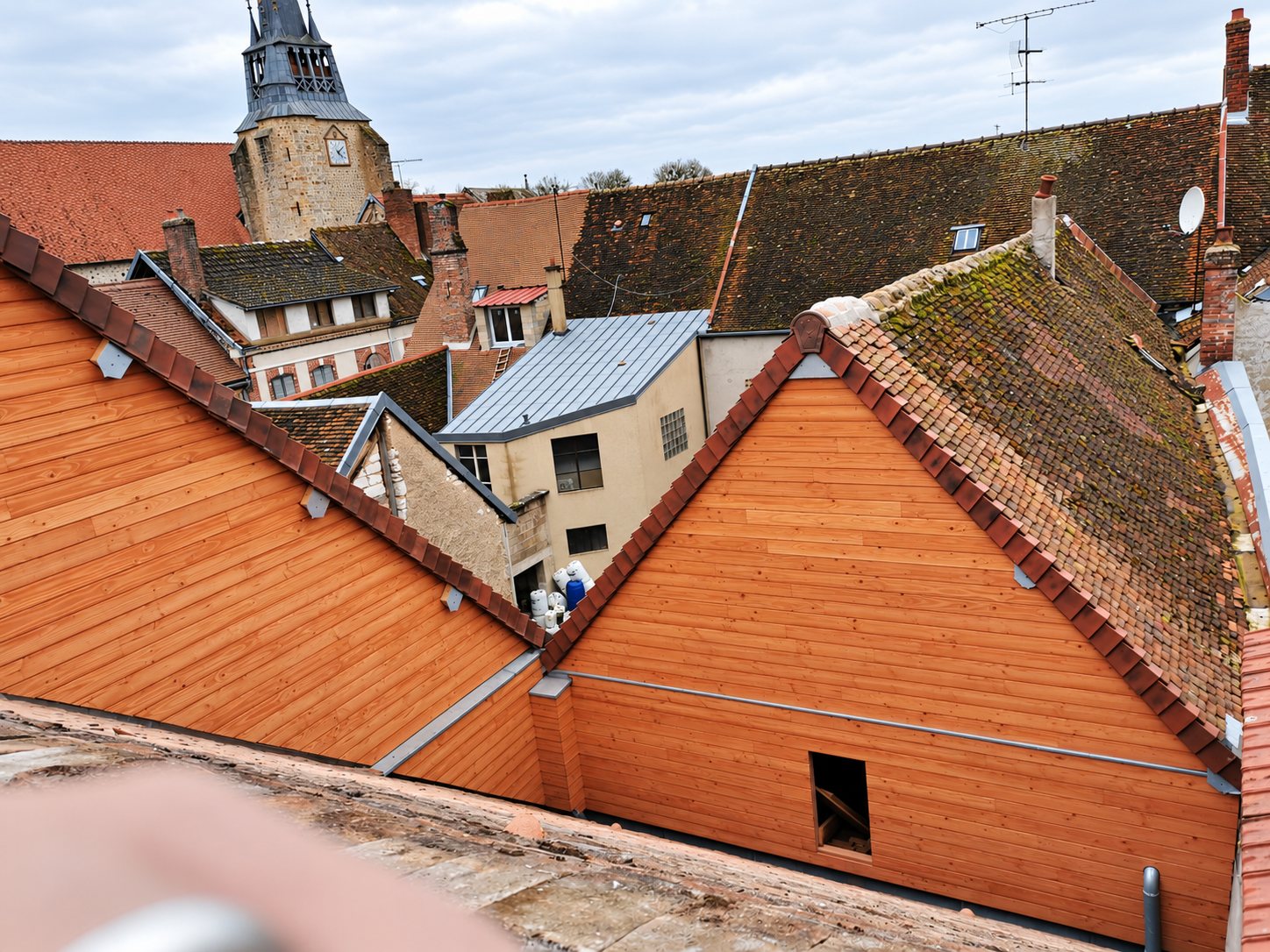 Vue depuis un toit sur deux pignons récemment habillés de bardage en bois clair, entourés de toitures anciennes en tuiles et d'un clocher en pierre.
