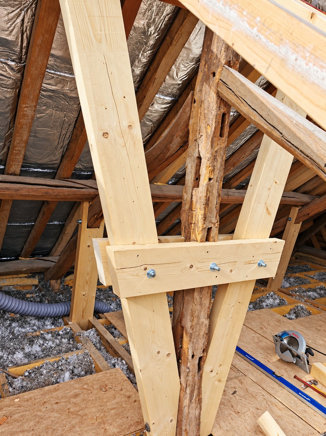Dans des combles isolés, de nouvelles poutres en bois clair sont fixées par des boulons de part et d'autre d'un poteau ancien abîmé, avec une scie circulaire posée sur le plancher.