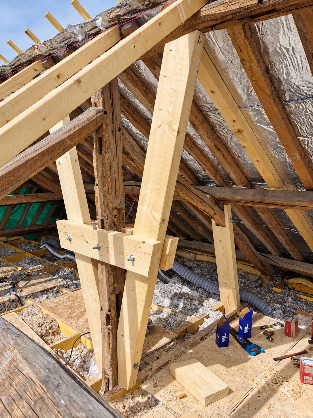 Dans des combles en chantier, de nouvelles poutres en bois clair boulonnées viennent renforcer un poteau ancien de la charpente.