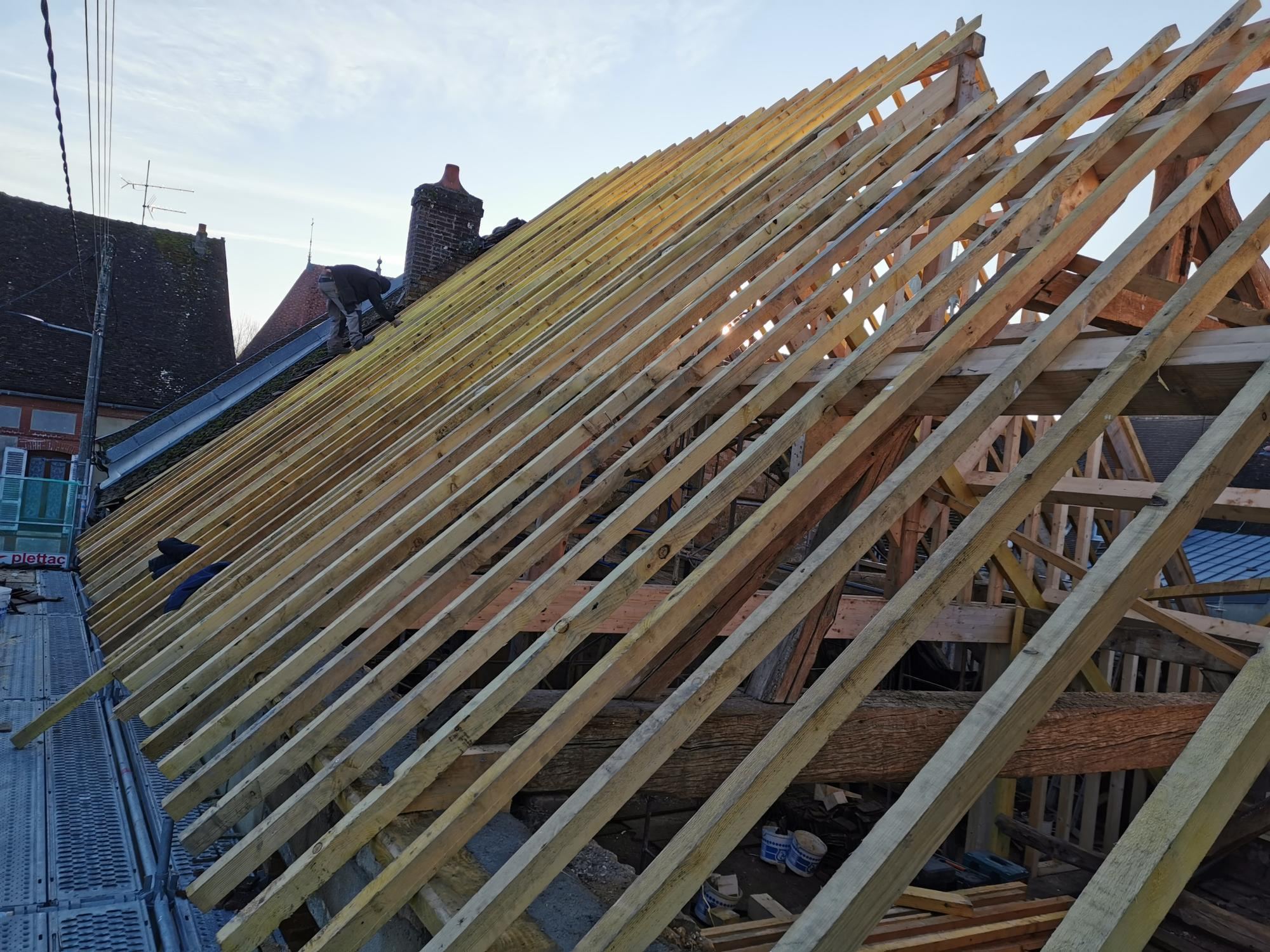 Une charpente ancienne en cours de rénovation, recouverte de nombreux chevrons en bois neufs, avec un ouvrier au travail sur le toit au coucher du soleil.