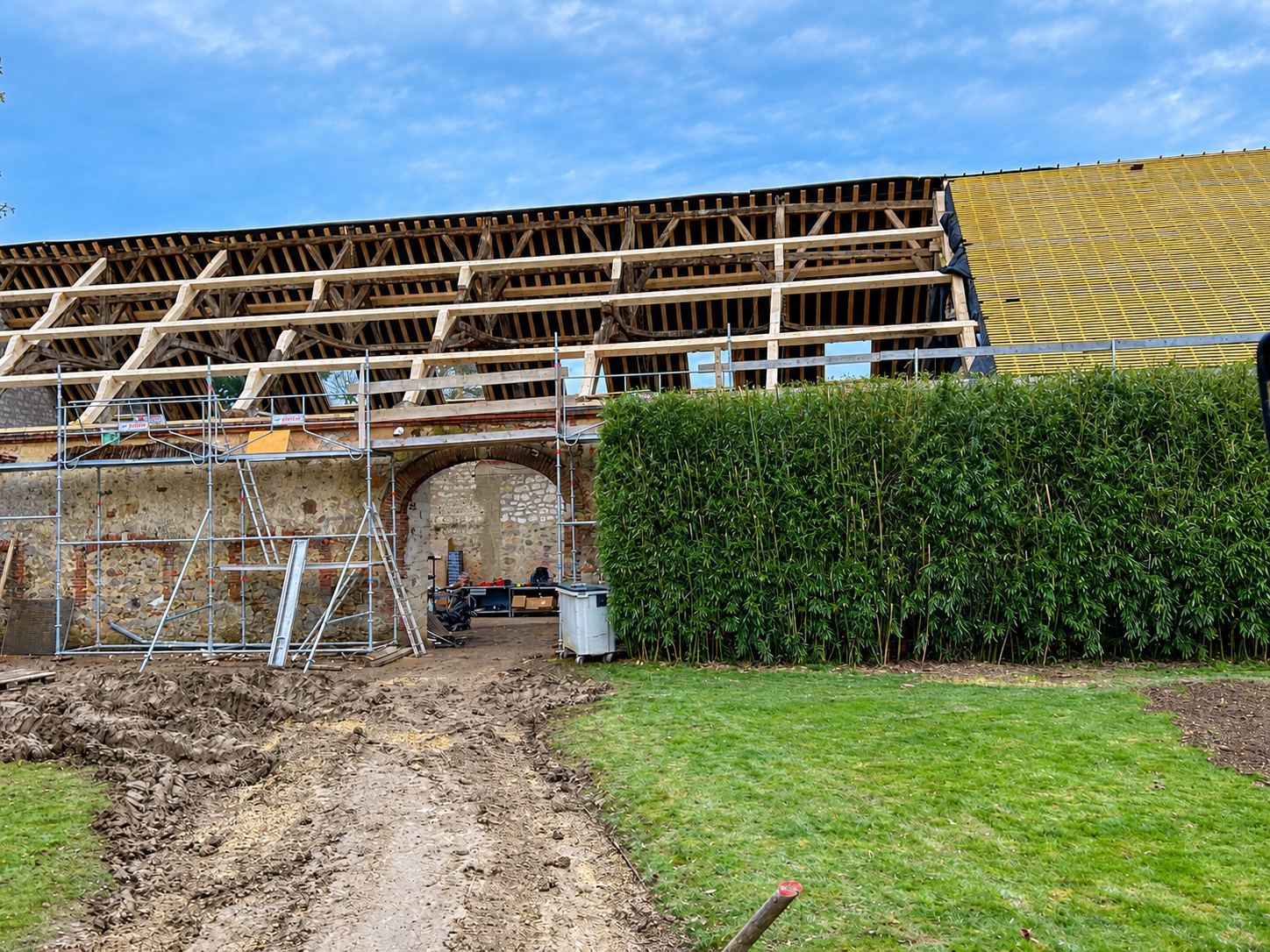 Chantier de rénovation d'une grange en pierre dont la charpente en bois est à nu, entourée d'un échafaudage, avec un chemin boueux et une haie de bambous au premier plan.