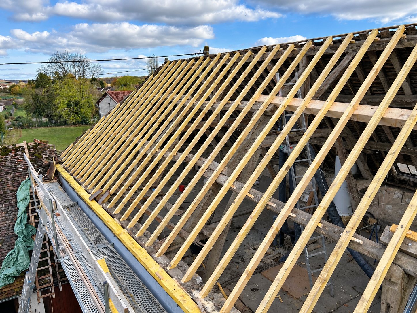 Vue depuis l'échafaudage d'une toiture en rénovation dont la vieille charpente est recouverte de nouveaux chevrons en bois clair, au-dessus d'un village.